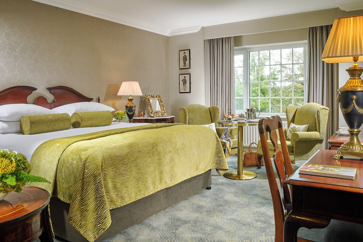 A hotel room with a large bed, a sitting area with a table and chairs, a lamp, and a view of the outdoors through a window.