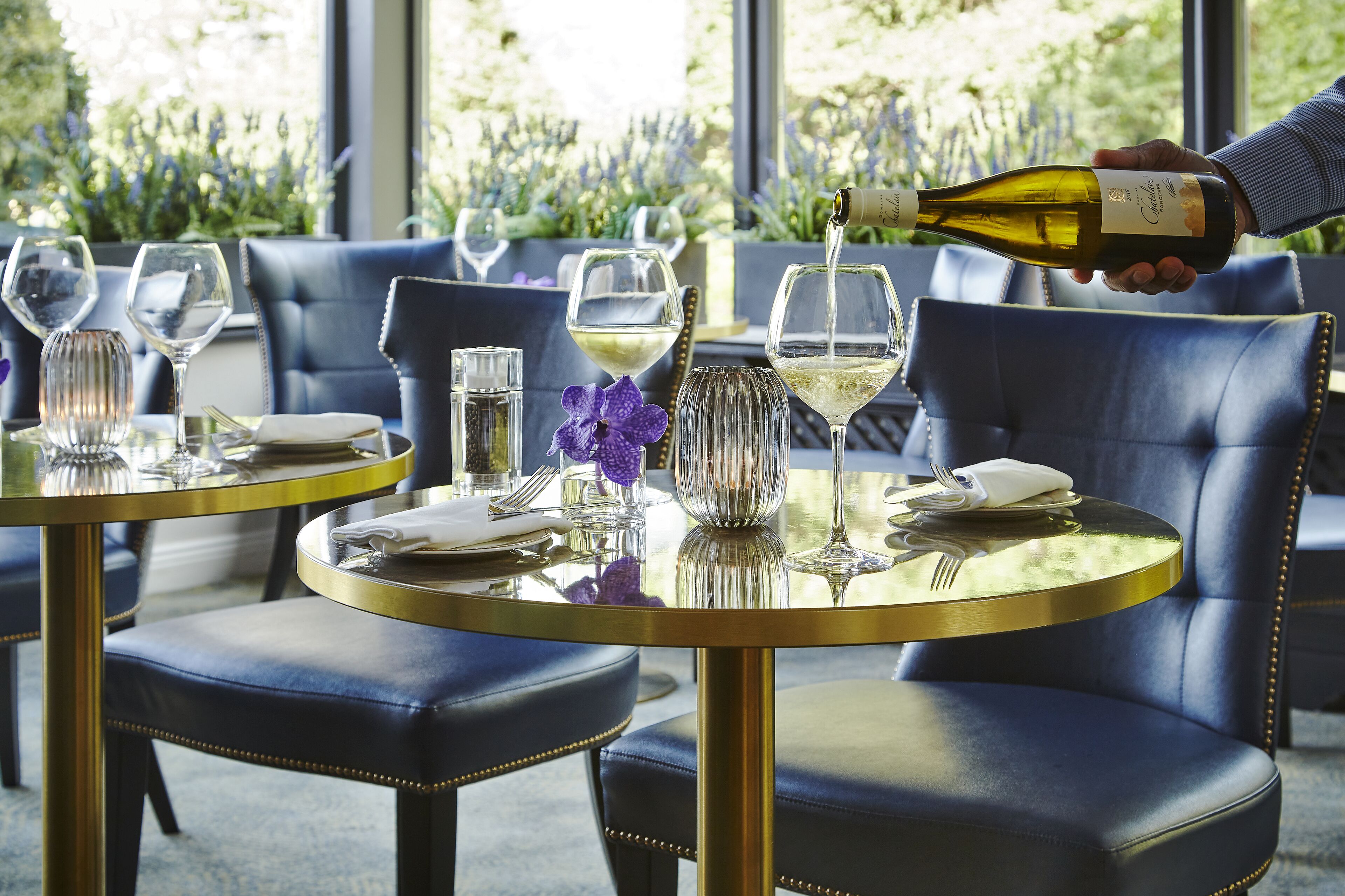 A dining area with round tables and blue leather chairs, a bottle of wine being poured into a glass, and a purple flower on the table.
