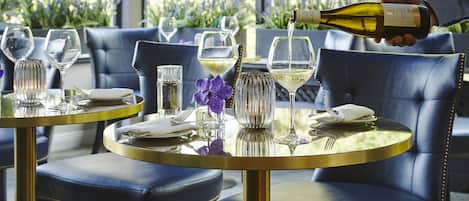 A dining area with round tables and blue leather chairs, a bottle of wine being poured into a glass, and a purple flower on the table.