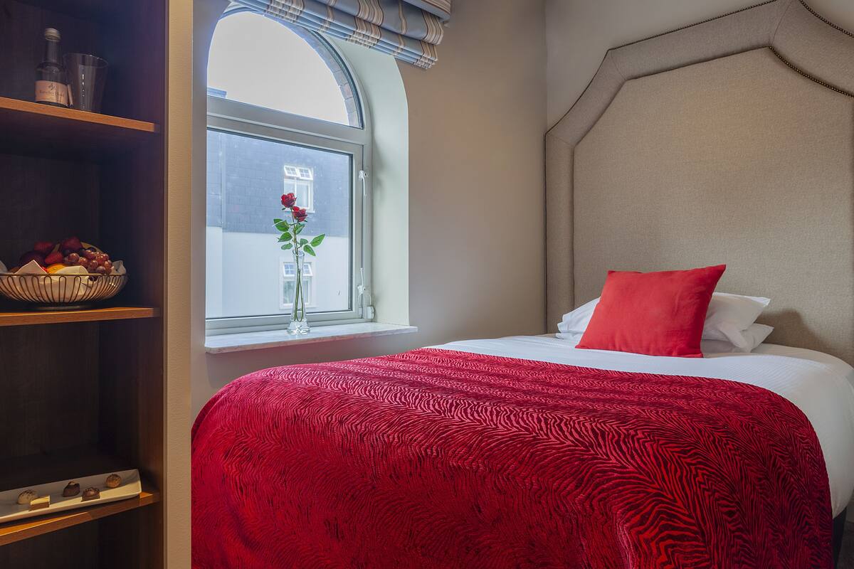 A hotel room with a large bed, a window with a view, a vase with flowers, and a shelf with snacks.