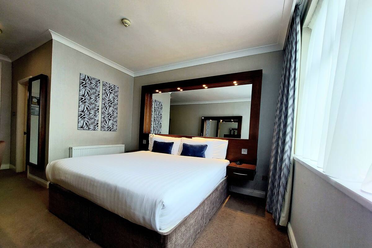 A hotel room with a large bed, a bedside table, a mirror, and a radiator.