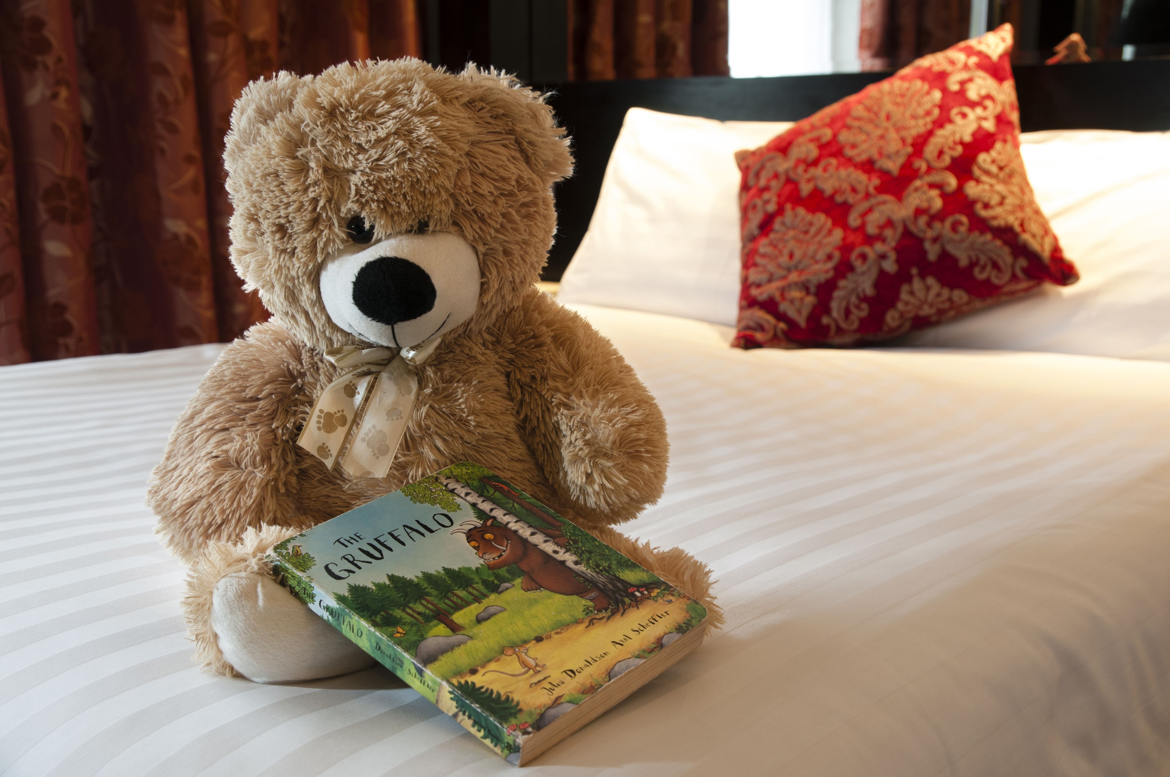 A plush teddy bear sitting on a bed with a book titled "The Gruffalo" and a red patterned pillow.