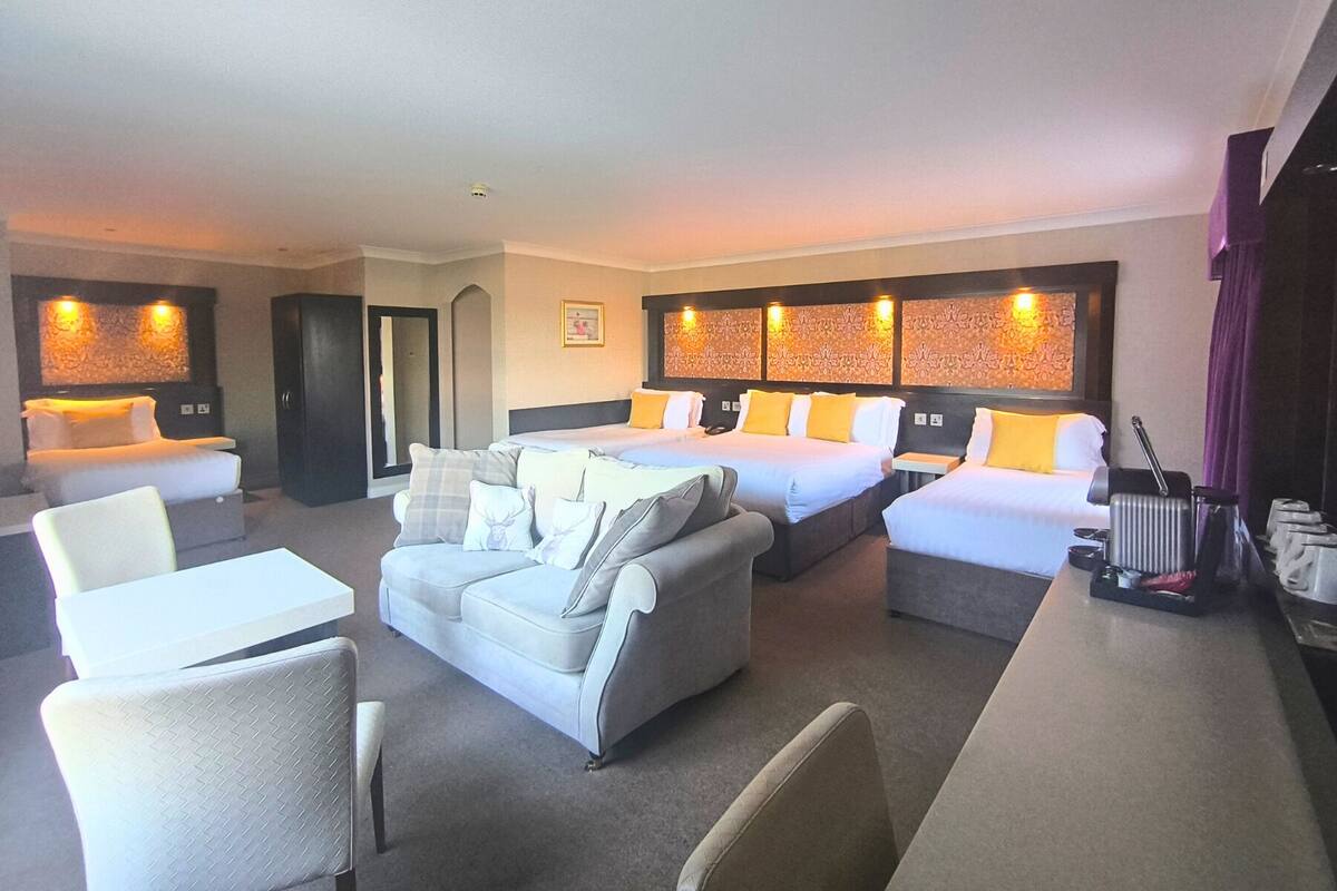 A hotel room with two single beds, a sofa, a dining table, and a small kitchen area.