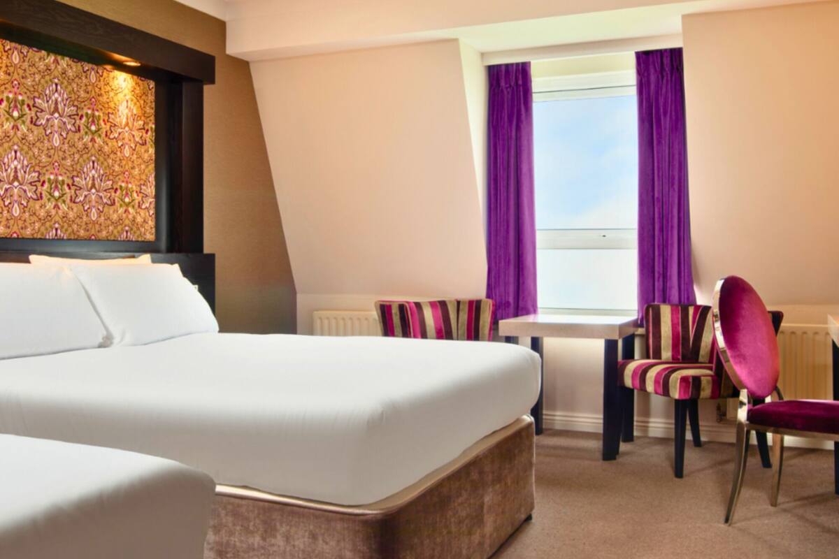 A hotel room with a large bed, a small table, two chairs, and a view of the sky through a window with purple curtains.