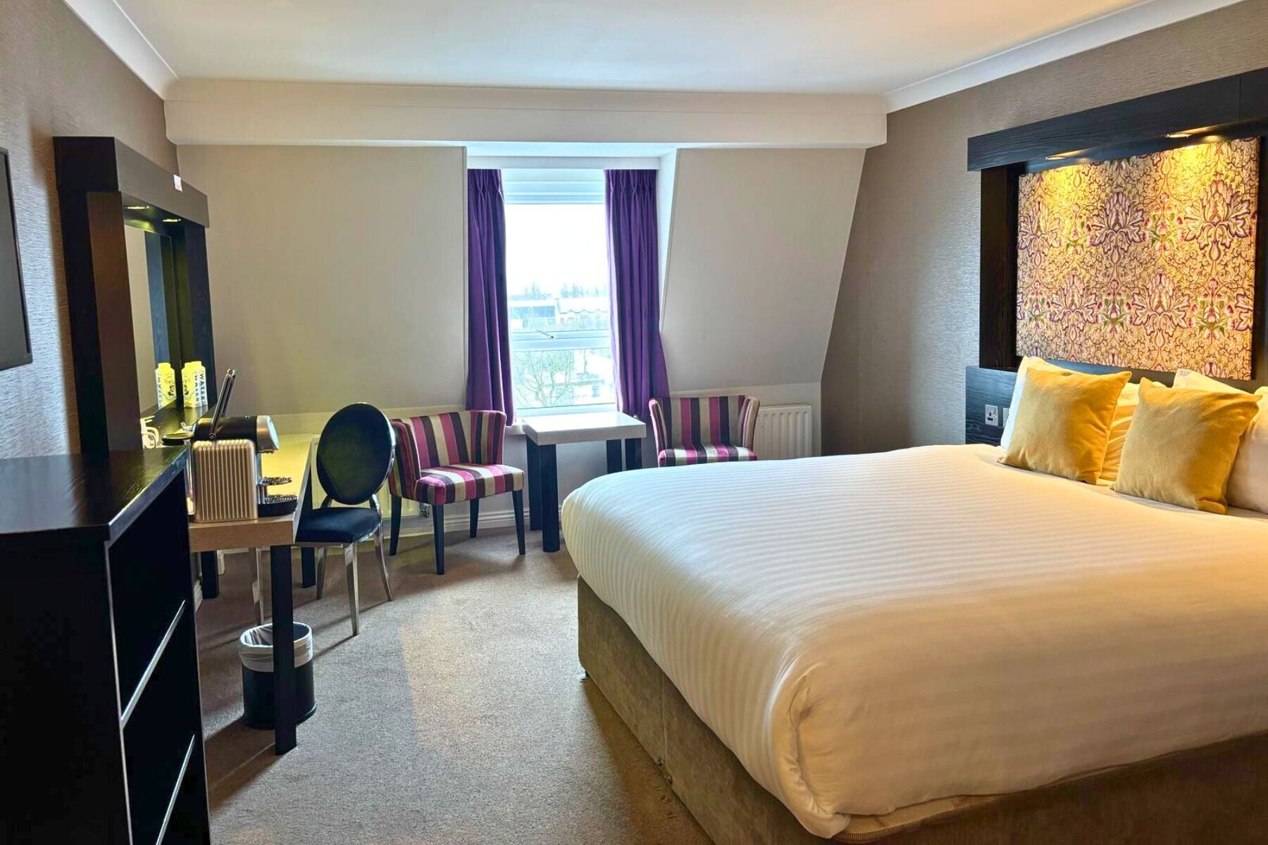 A hotel room with a bed, a desk, a chair, a TV, and a window with curtains.