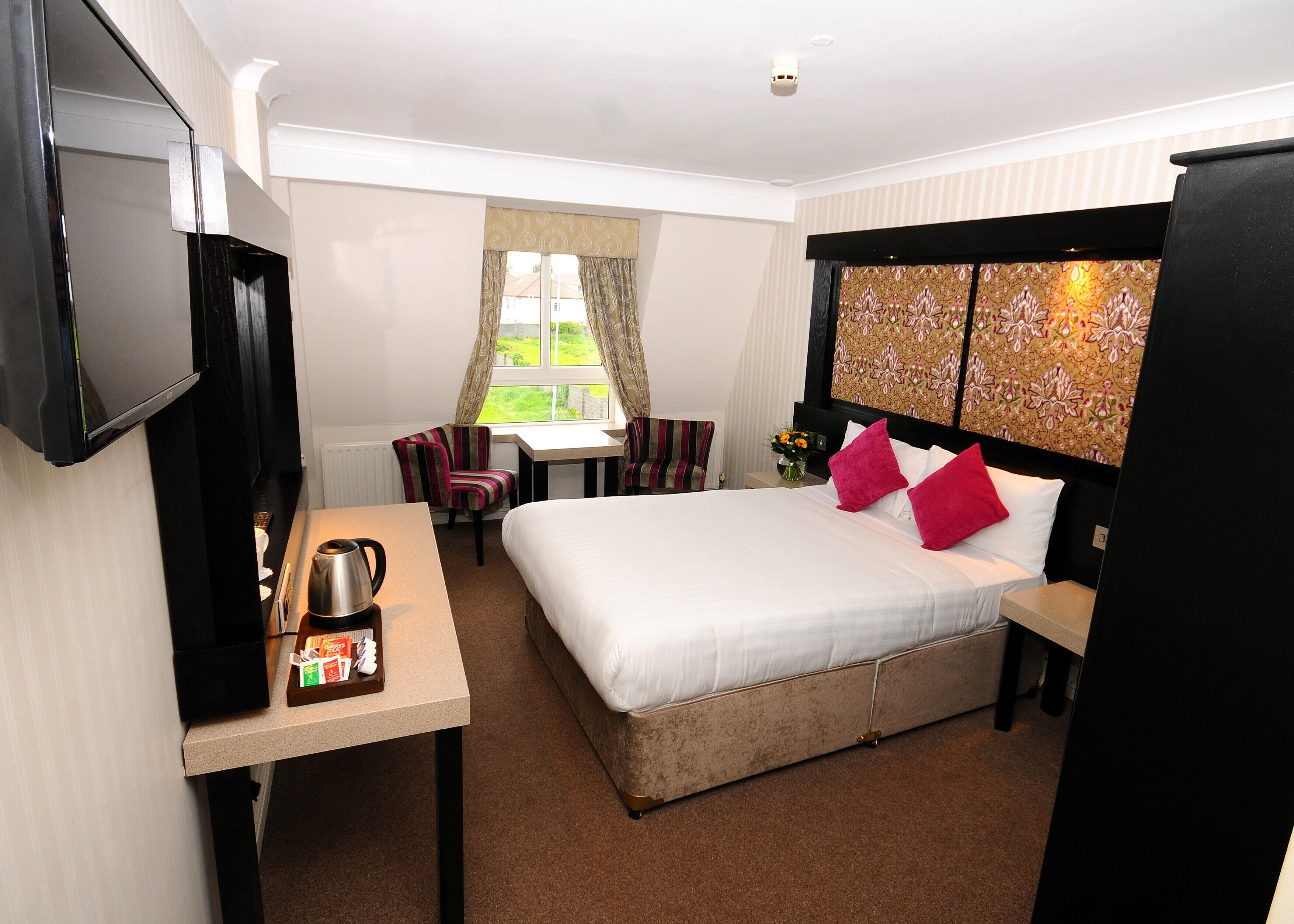 A hotel room with a bed, a desk with a coffee maker, a TV, and a window with curtains.