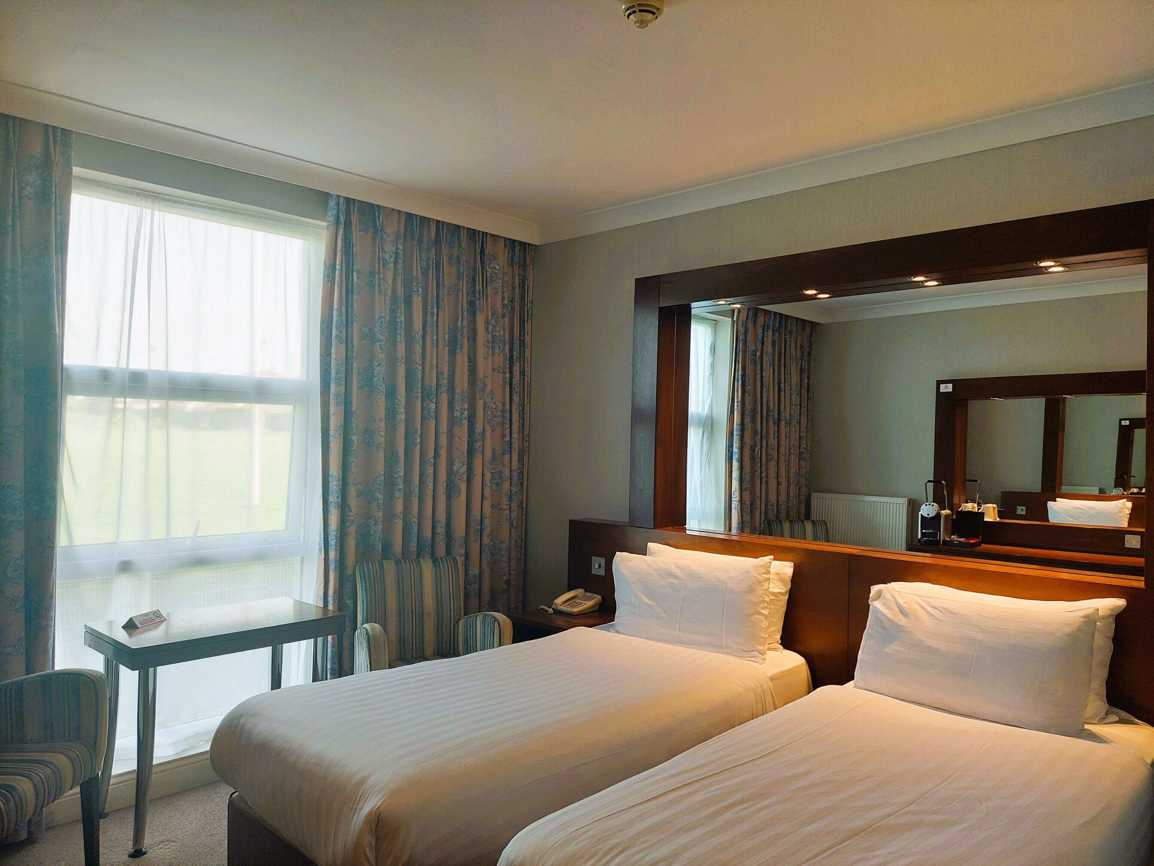 A hotel room with two beds, a desk, a chair, a large mirror, and a window with curtains.