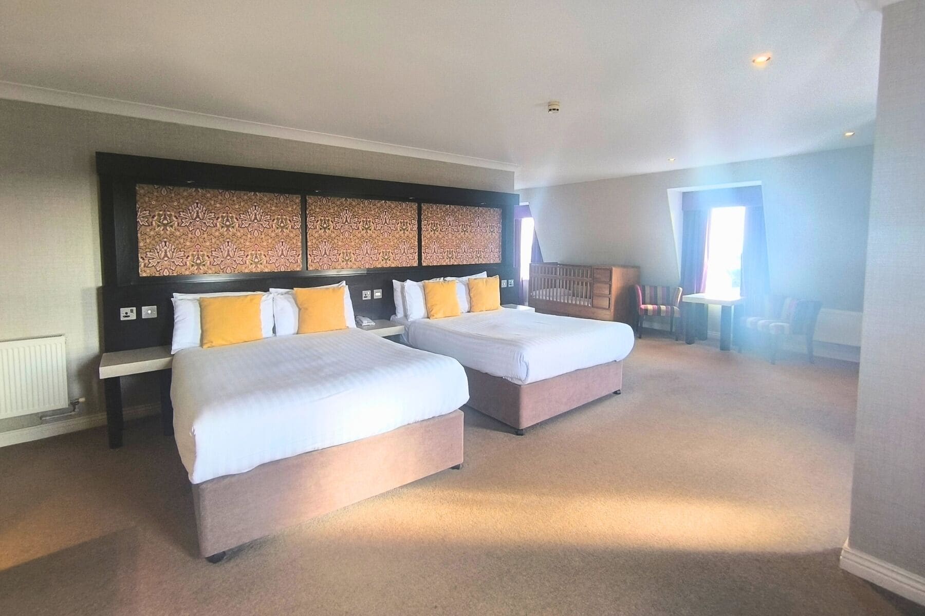 Quarto de hotel com duas camas, uma cabeceira decorativa grande, uma mesinha e uma cadeira.