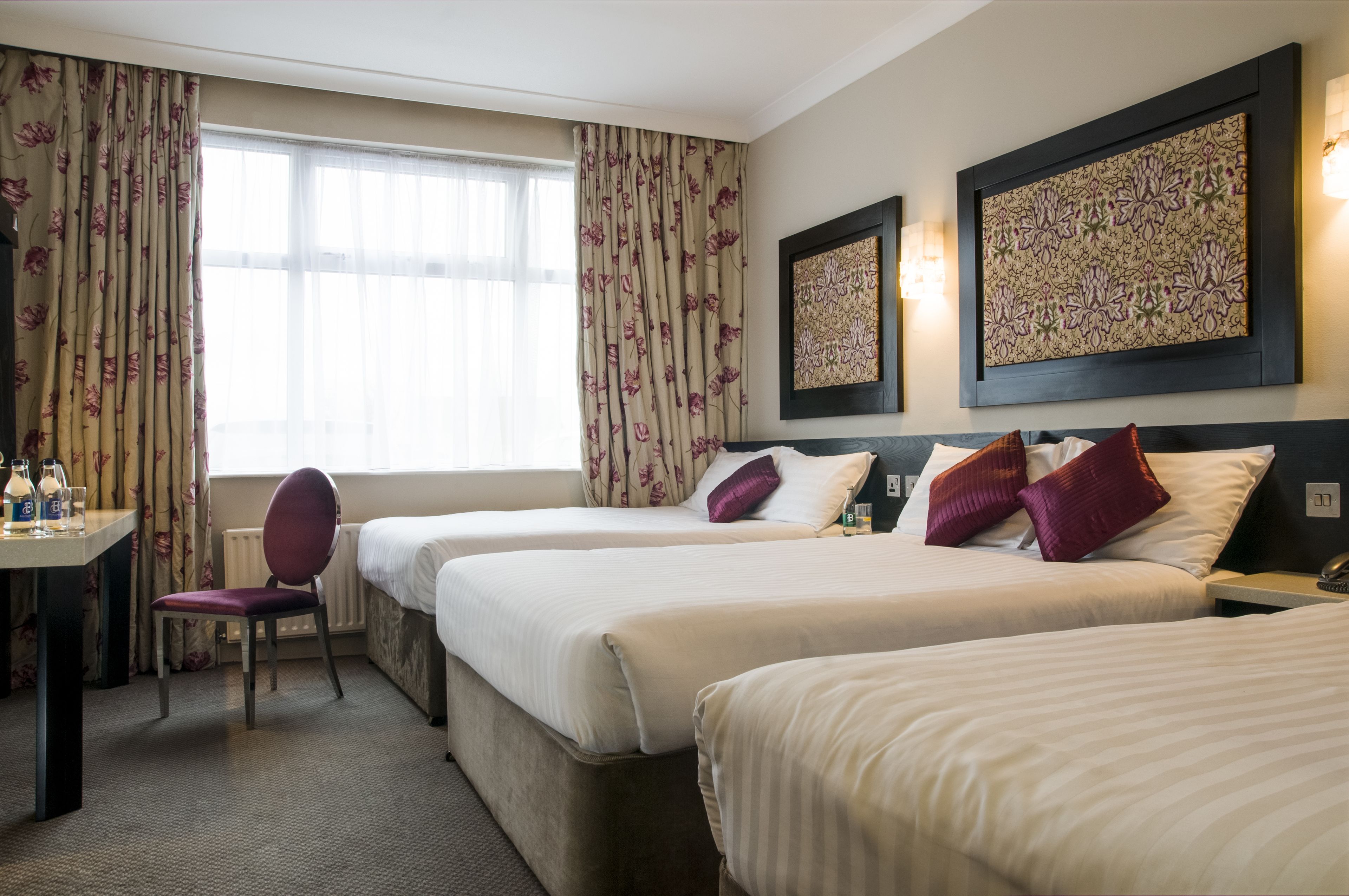 A hotel room with two beds, a desk, and a chair. There are floral curtains, wall art, and a view of the outdoors.