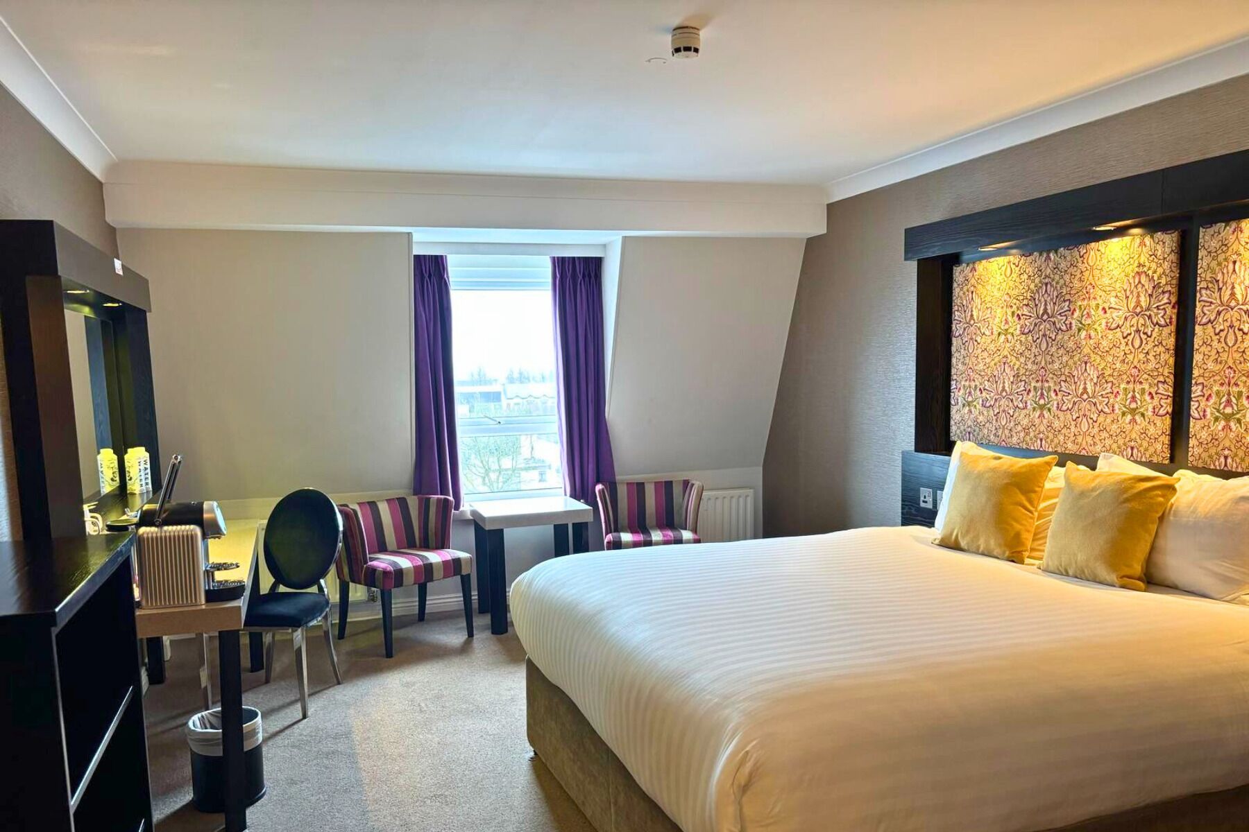 Habitación de hotel con una cama grande, un escritorio con silla, una mesita y una ventana con cortinas.