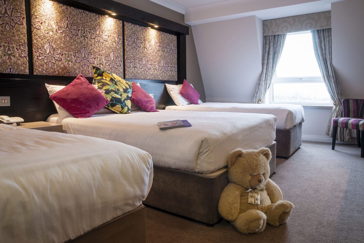 A hotel room with two beds, a large decorative headboard, a teddy bear, and a window with curtains.