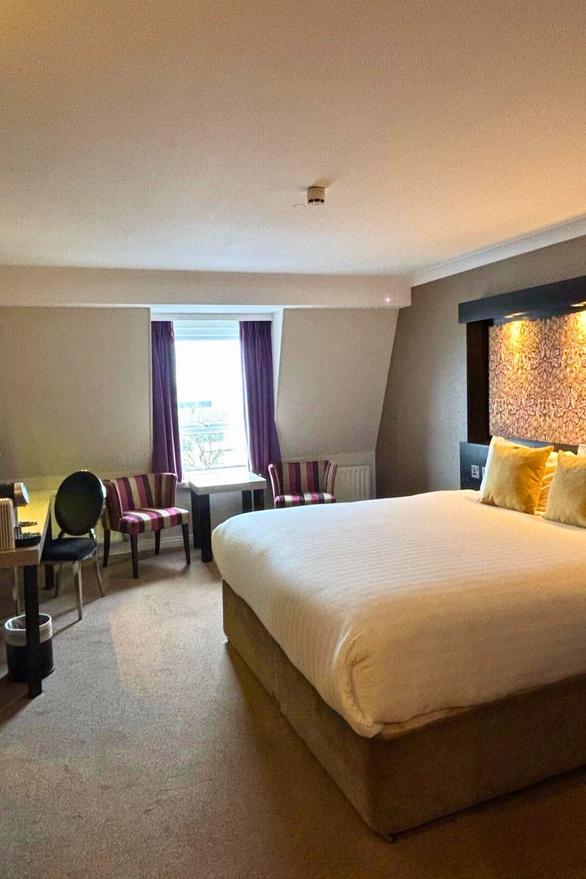 A hotel room with a large bed, a desk with a chair, and a window with curtains.