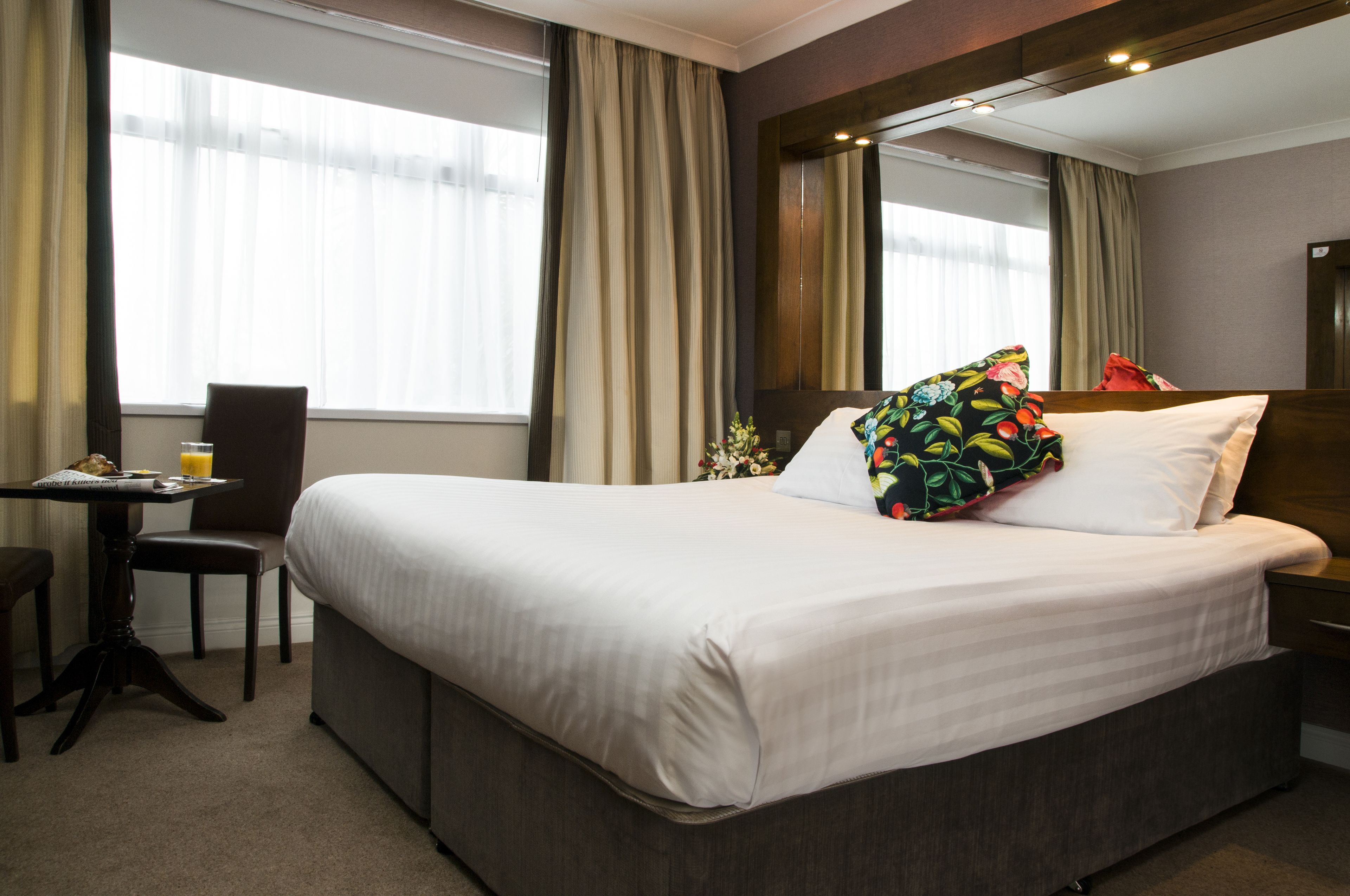 A hotel room with a large bed, a desk with a chair, a small table with a glass of orange juice, and a flower arrangement.