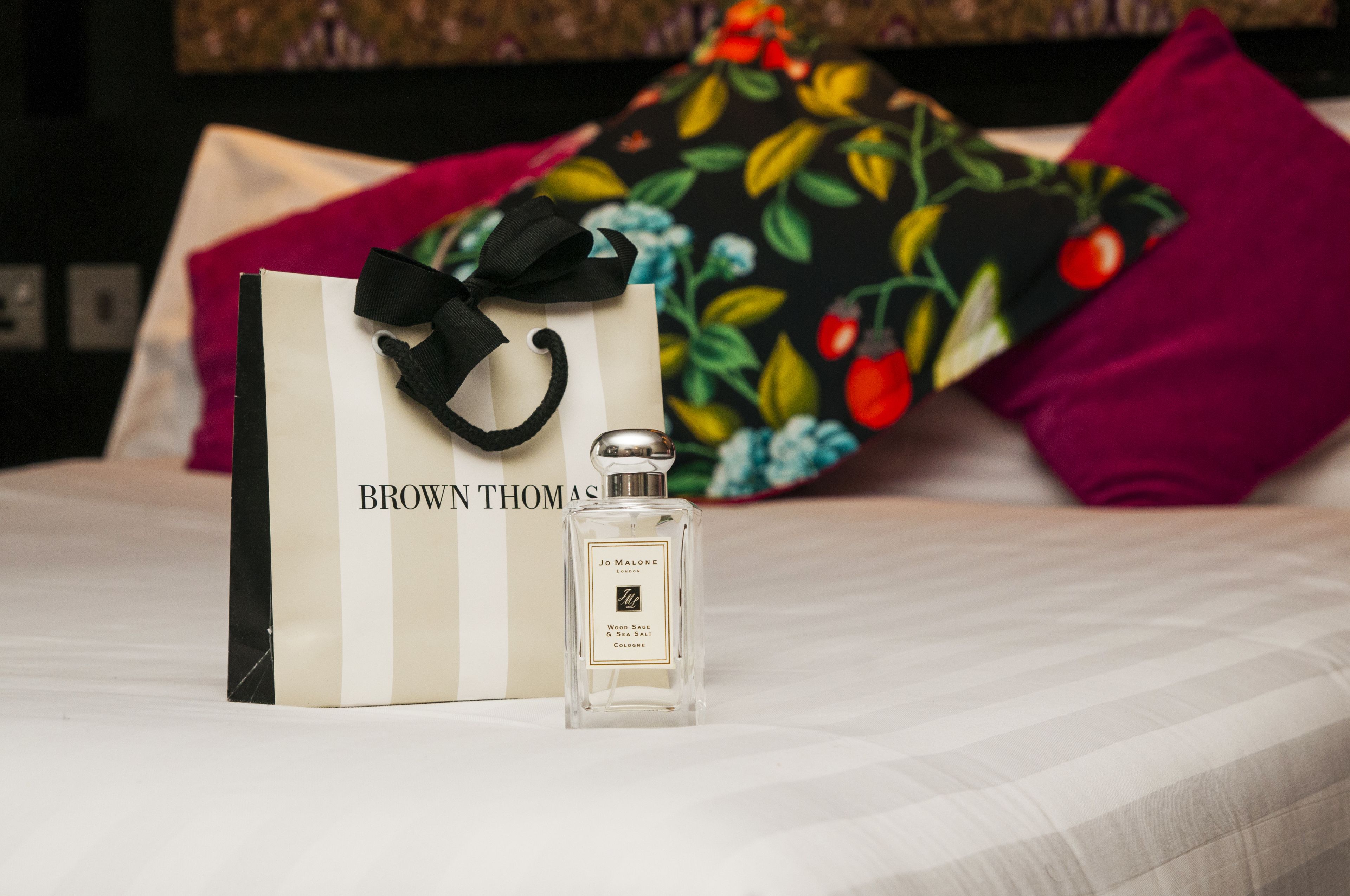 Una botella de colonia Jo Malone y una bolsa de regalo de Brown Thomas sobre una cama con un cojín floral y un cojín rojo al fondo.