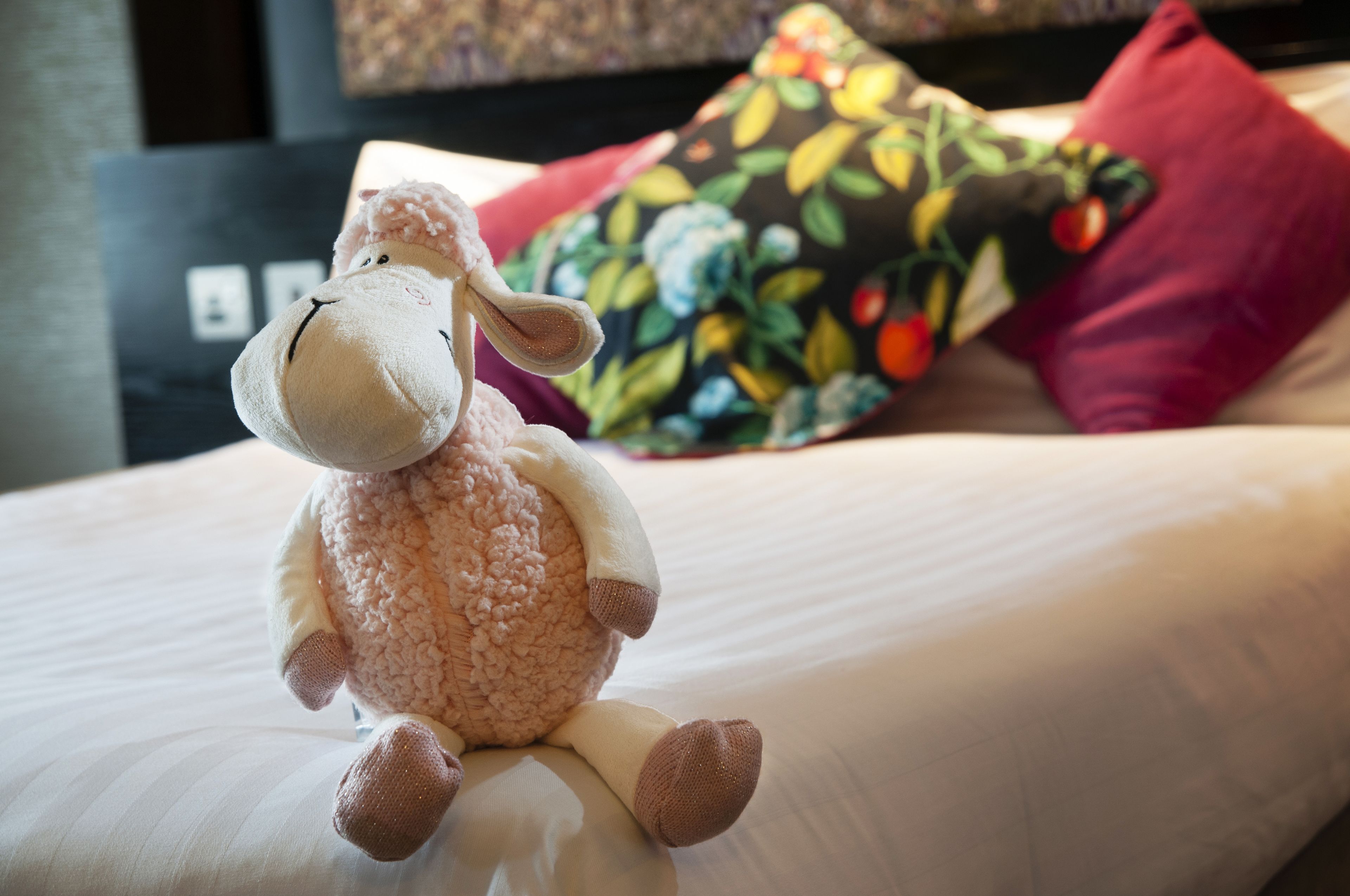 A plush toy on a bed with a floral pillow and a red pillow in the background.