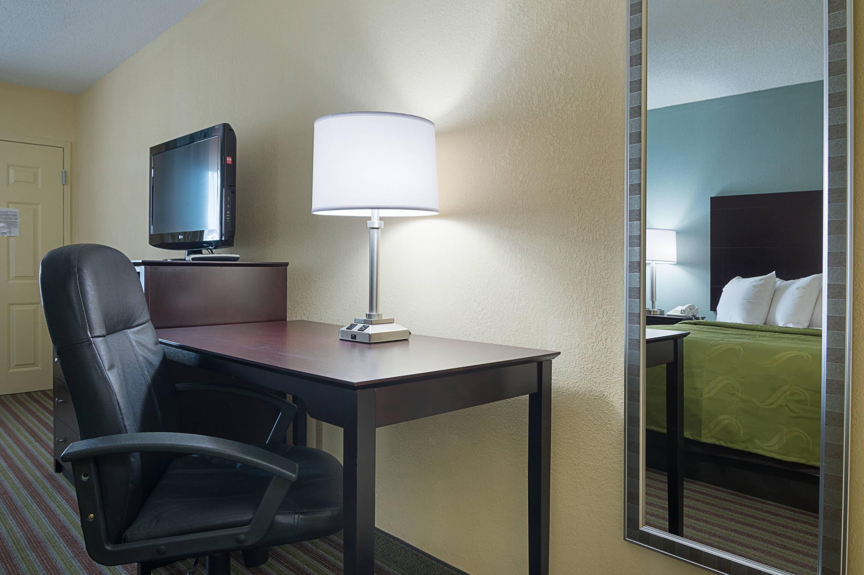 A hotel room with a desk, chair, TV, lamp, and a bed with a green bedspread.