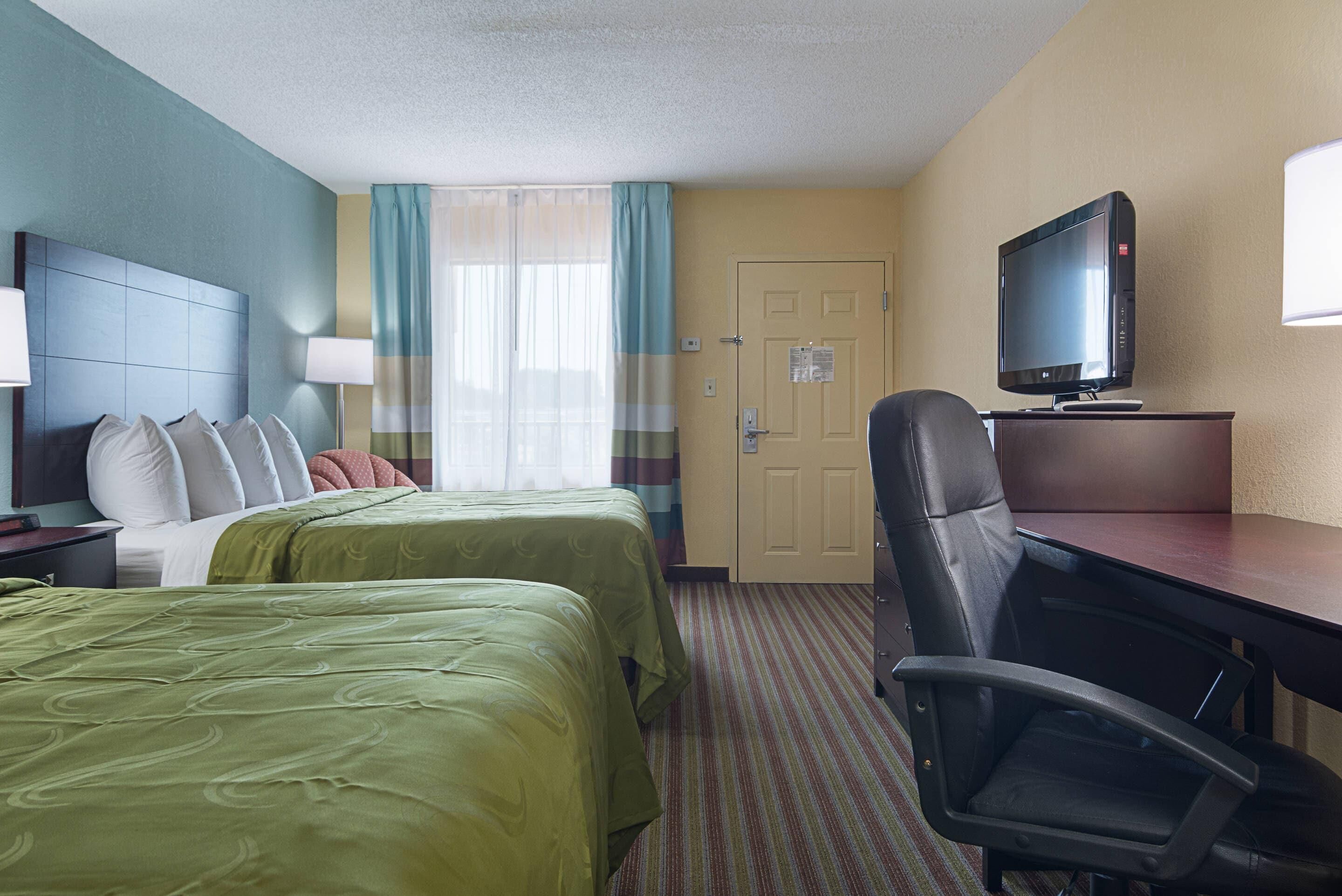 A hotel room with two beds, a desk, a chair, a television, and a door.