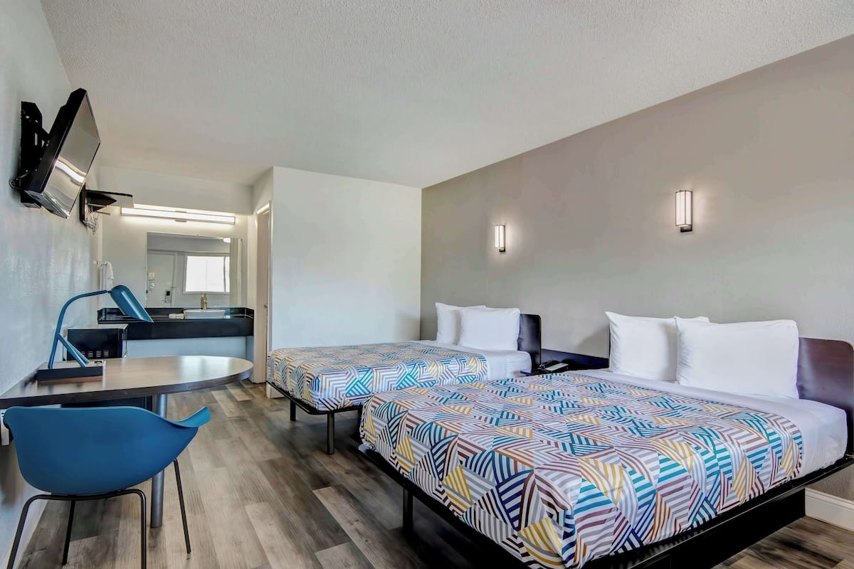 A hotel room with two beds, a desk, a chair, a TV, and a bathroom visible in the background.