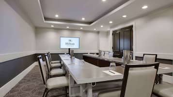 A conference room with a long table, chairs, a projector screen displaying the Hilton logo, and a carpeted floor.