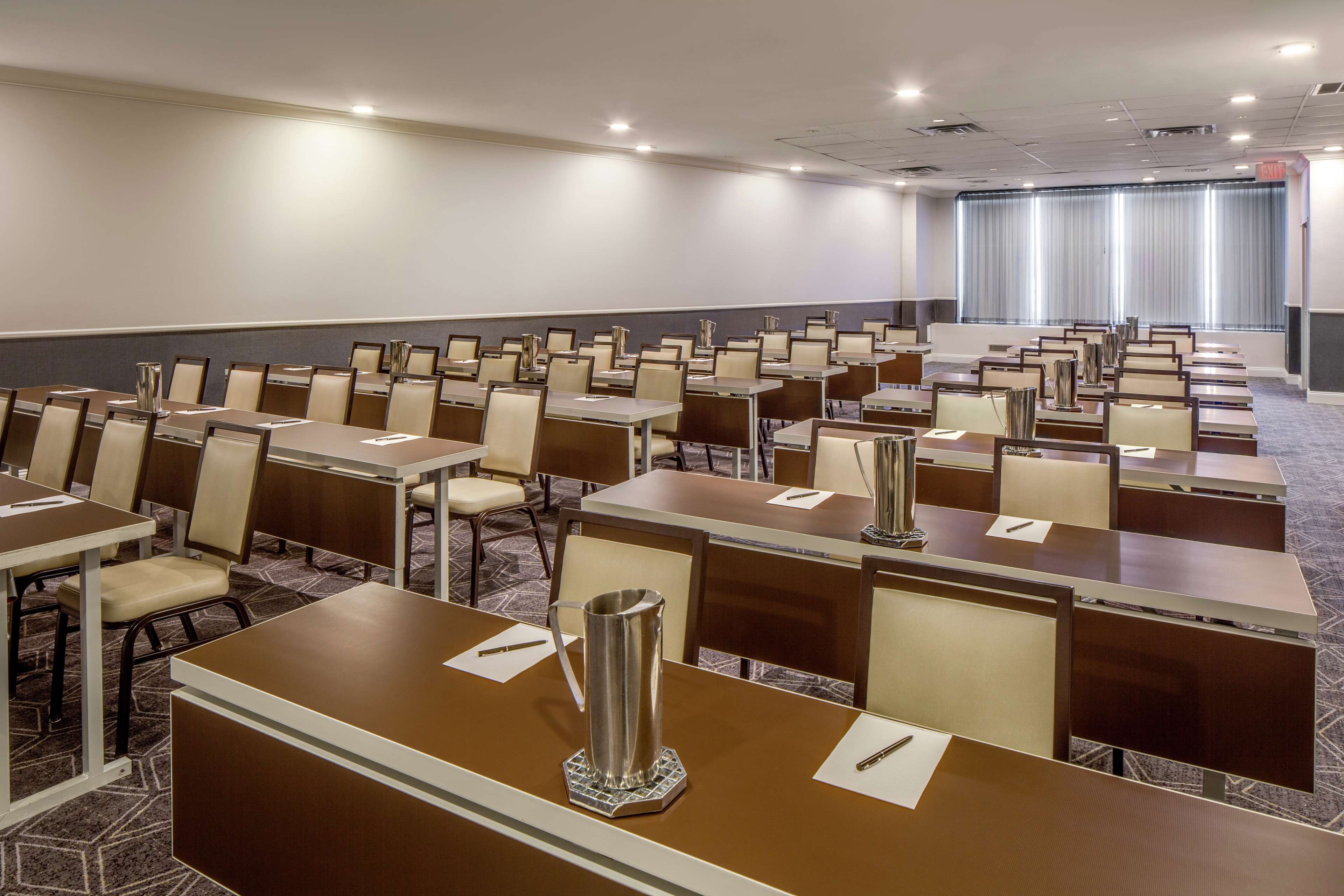 A conference room with rows of tables and chairs, a large window with curtains, and a carpeted floor.