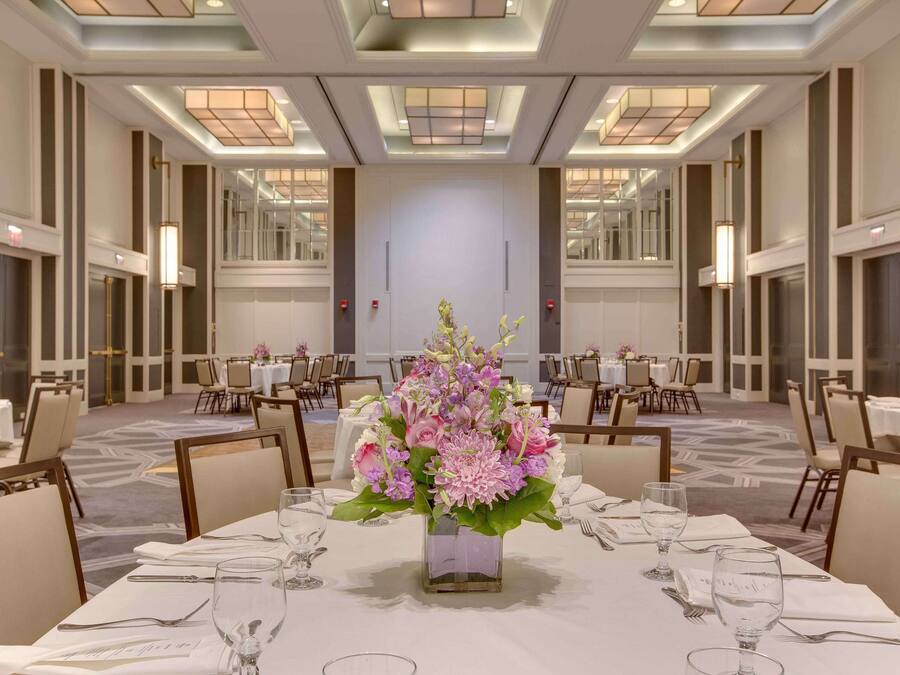 Uma ampla sala de banquetes com mesas redondas, cada uma com toalhas brancas, talheres e copos de vidro transparente. Um grande arranjo floral decora a mesa central. O espaço possui pé-direito alto com iluminação embutida e um espelho na parede do fundo.