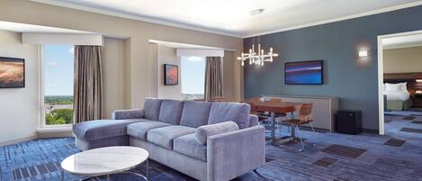 A modern hotel room with a grey sectional sofa, a round coffee table, a dining table with chairs, a large window with curtains, and a city view.
