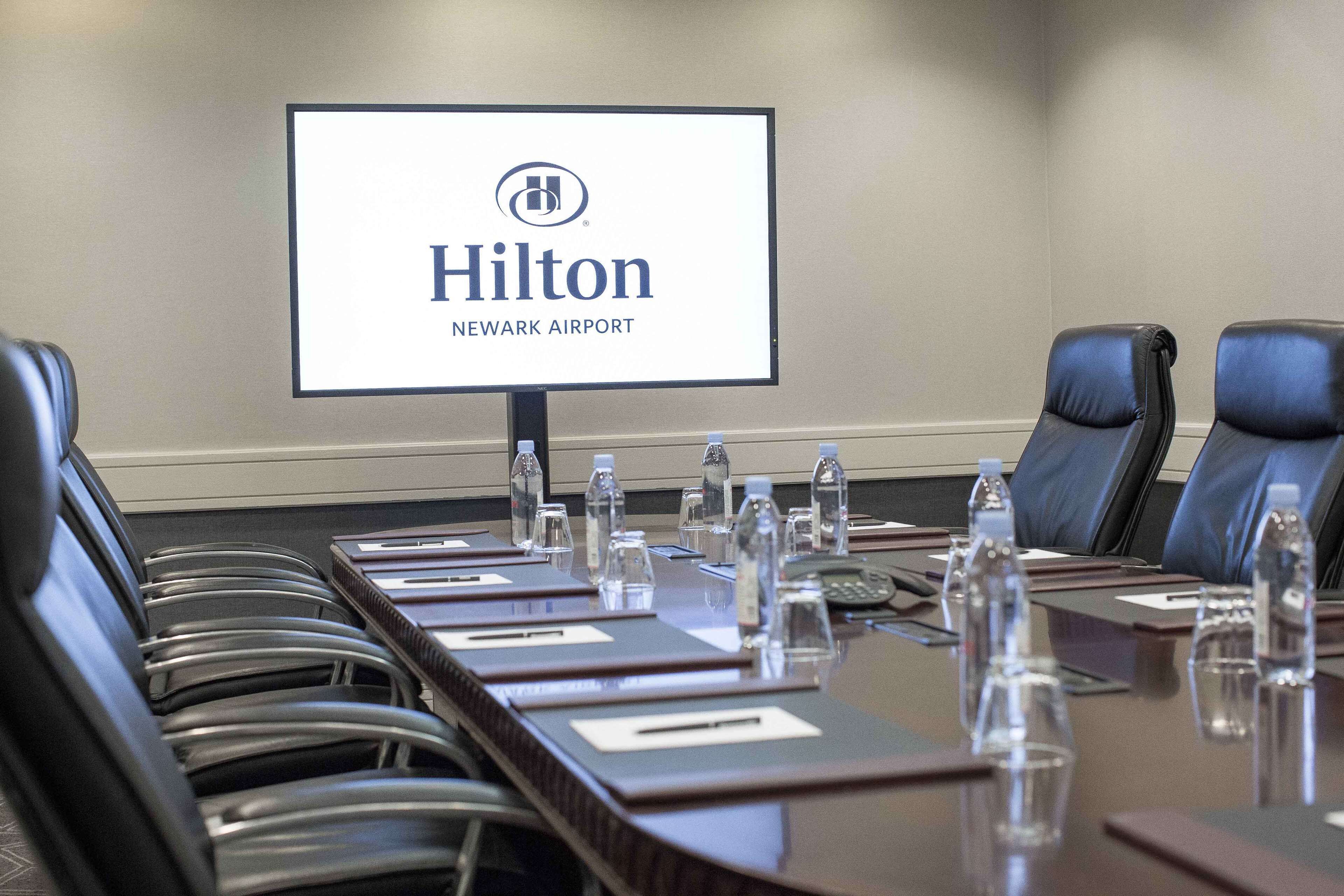 A conference room with a long table, black leather chairs, water bottles, and a screen displaying the Hilton Newark Airport logo.