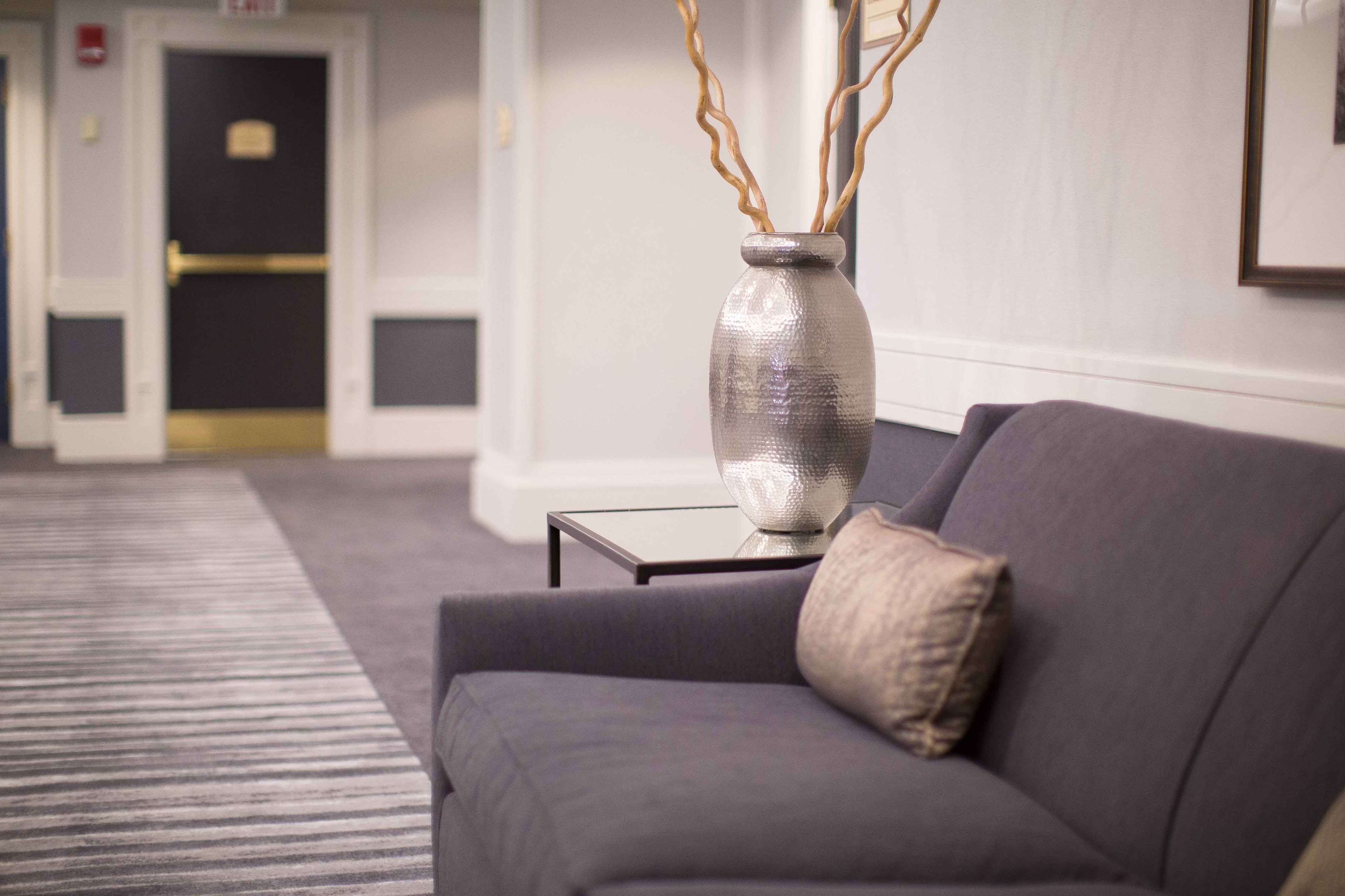A modern hotel lobby with a grey sofa, a metallic vase with branches, and a framed picture on the wall.