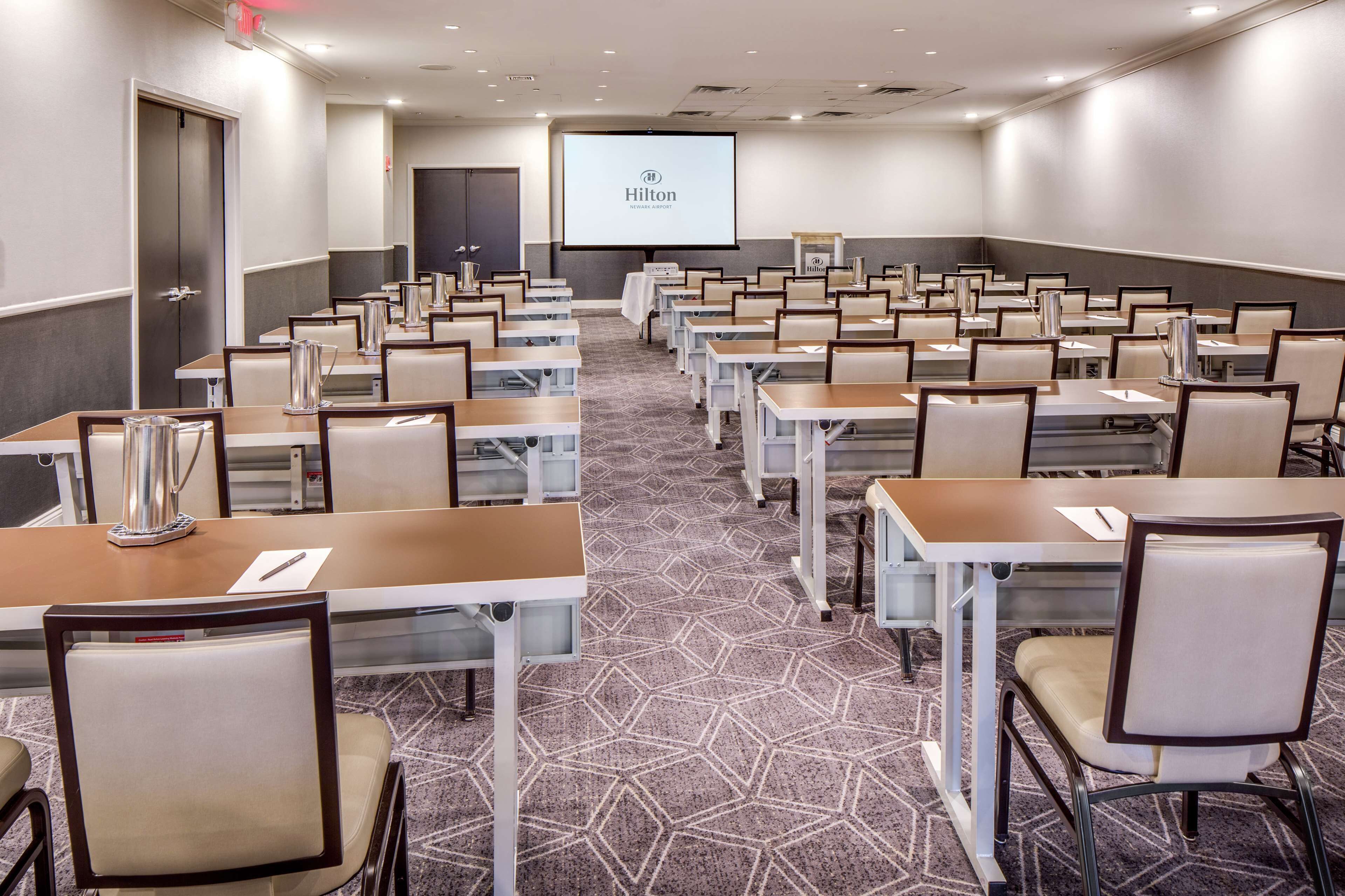A conference room with multiple tables and chairs, a projector screen displaying the Hilton logo, and a carpeted floor.
