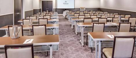 A conference room with multiple tables and chairs, a projector screen displaying the Hilton logo, and a carpeted floor.