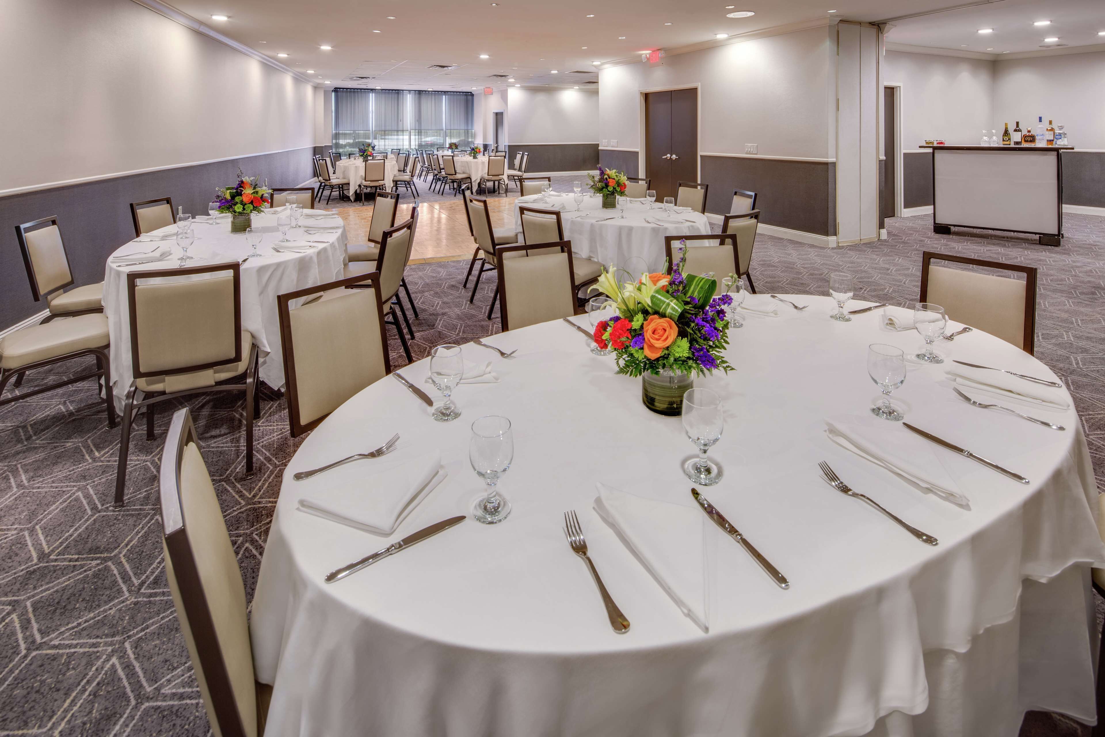 A conference room with round tables set for dining, each with white tablecloths, chairs, and floral centerpieces.