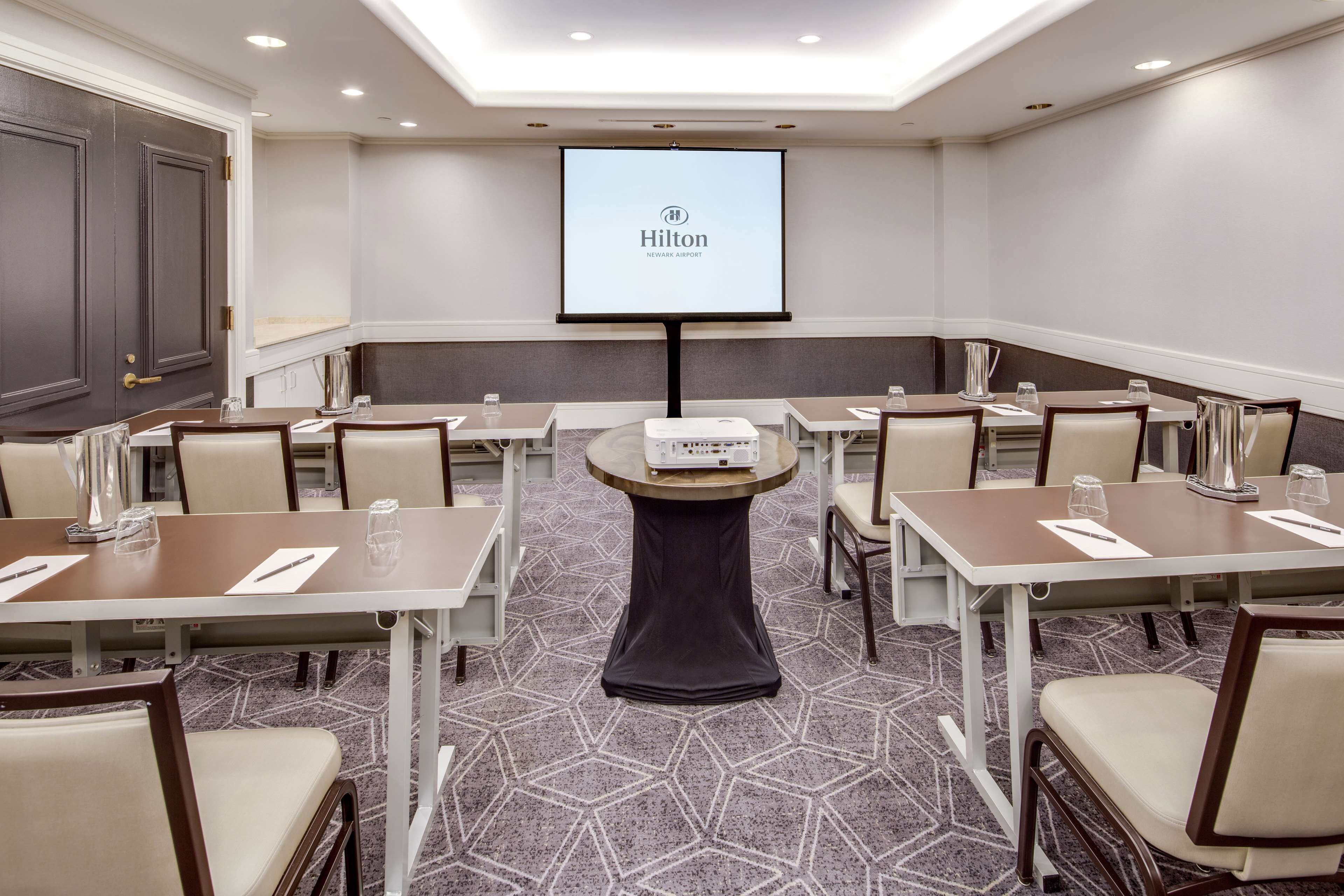 Une salle de conférence dotée d’un grand écran affichant le logo de Hilton, de plusieurs tables avec des chaises et d’un sol recouvert de moquette.