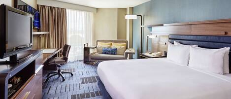 A hotel room with a large bed, a desk with a flat-screen TV, a chair, a bedside table with a lamp, and a window with curtains.