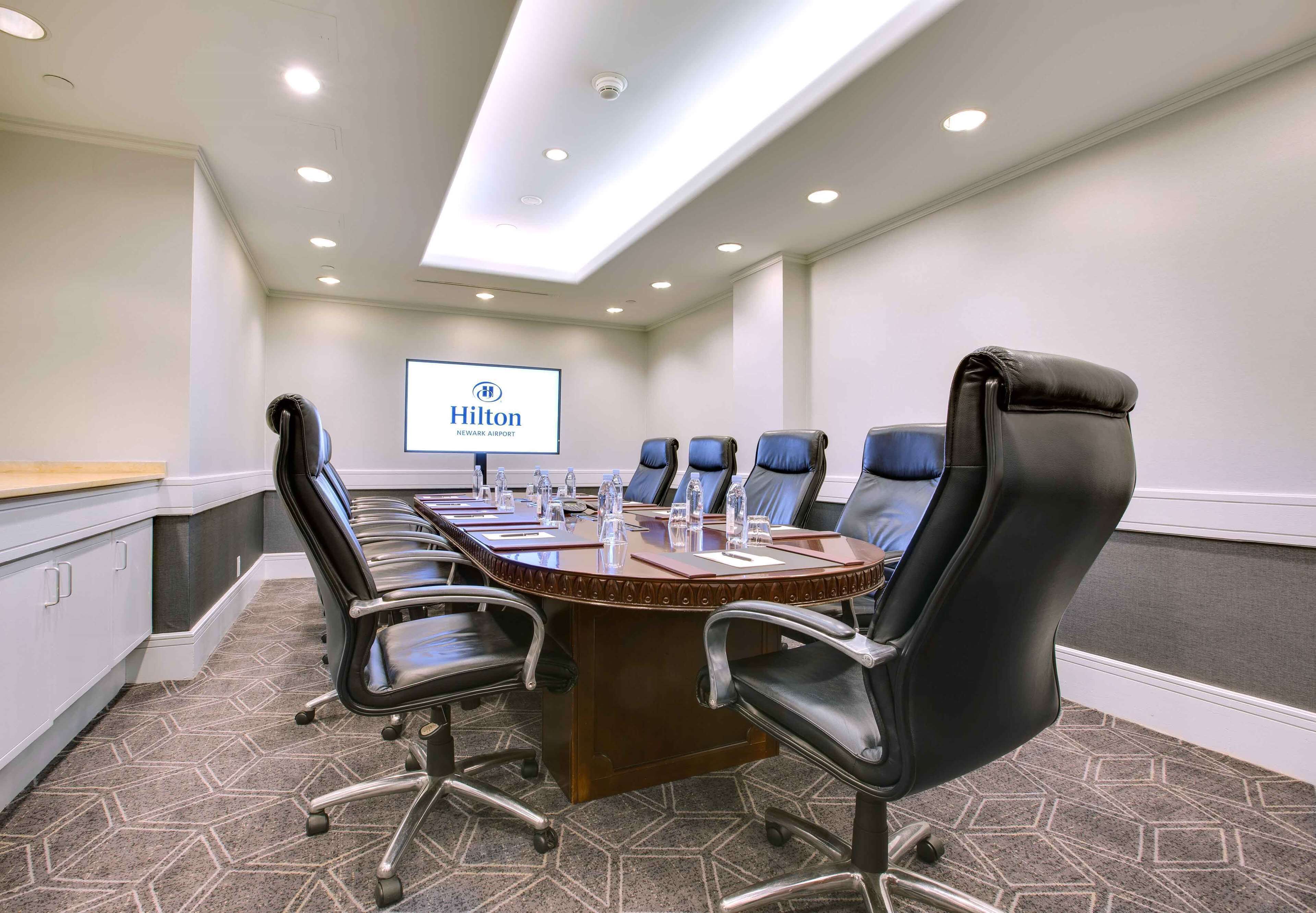 A conference room with a long table, black chairs, and a screen displaying the Hilton logo.