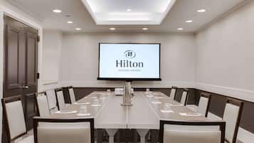 Un salón de conferencias con una larga mesa, sillas, una pantalla de proyector mostrando 'Hilton Newark Airport' e iluminación empotrada en el techo.