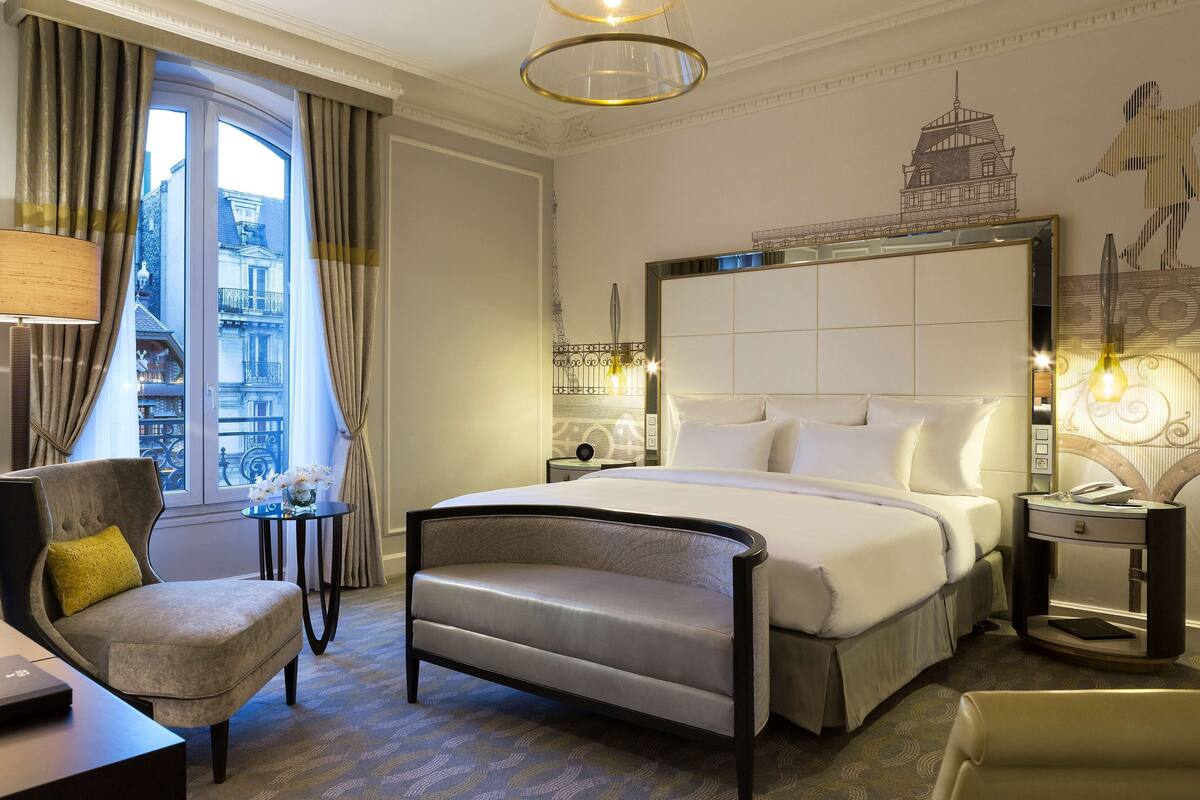 Habitación de hotel con una cama grande, una silla, un escritorio, una mesita de noche y vistas a un edificio a través de una ventana.