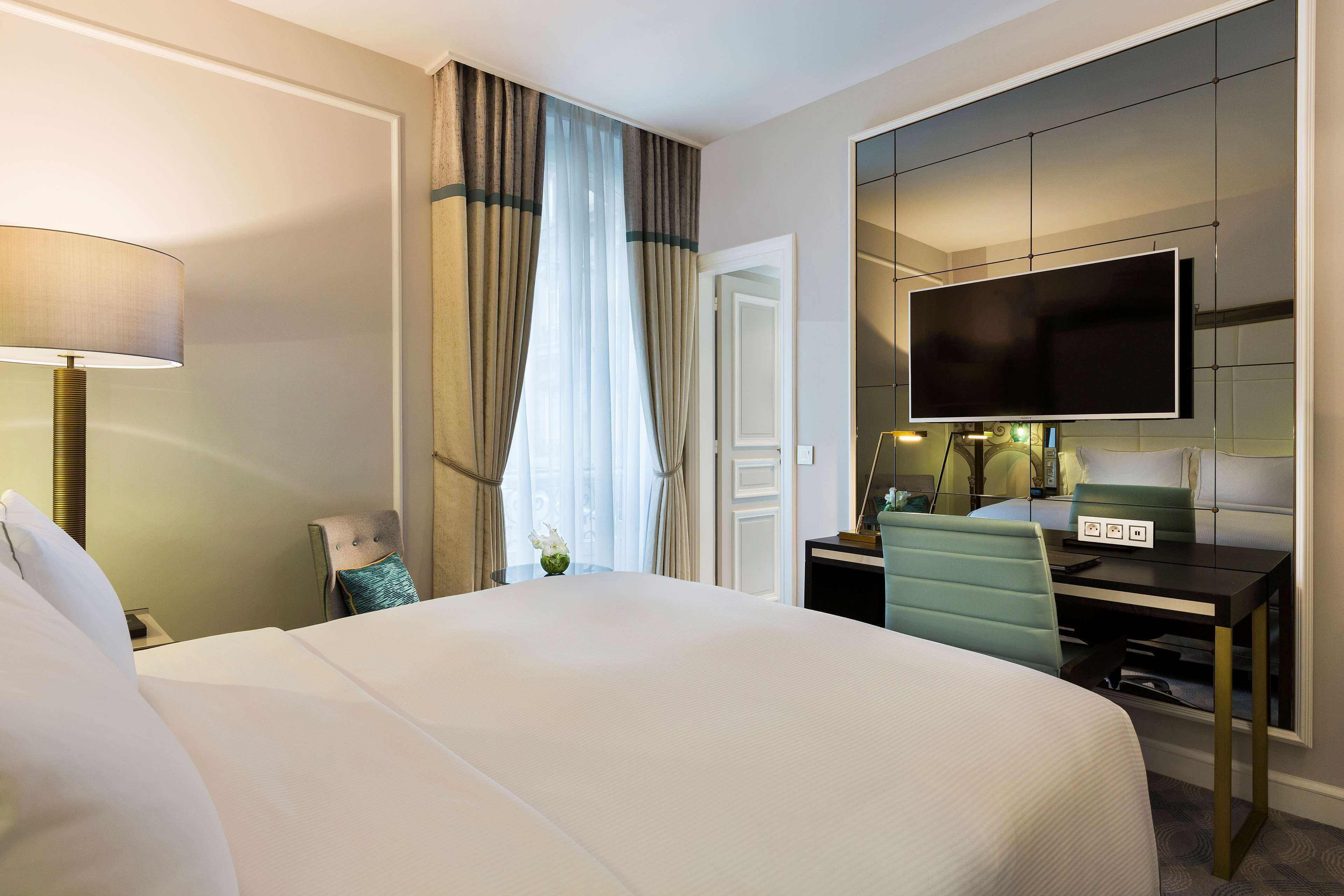 A hotel room with a large bed, a desk with a flat-screen TV, a chair, and a lamp.