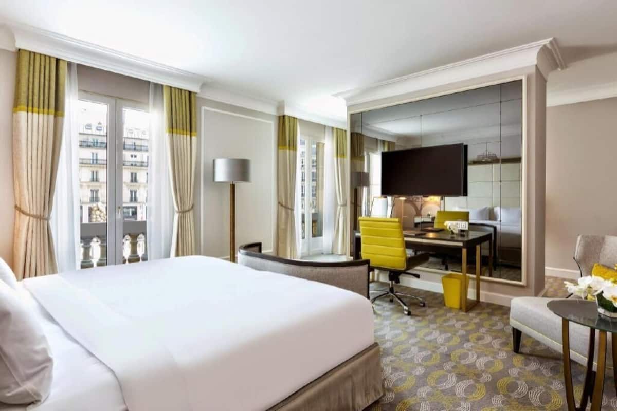 Habitación de hotel con una cama grande, un escritorio con una silla, un televisor y vistas a edificios a través de la ventana.