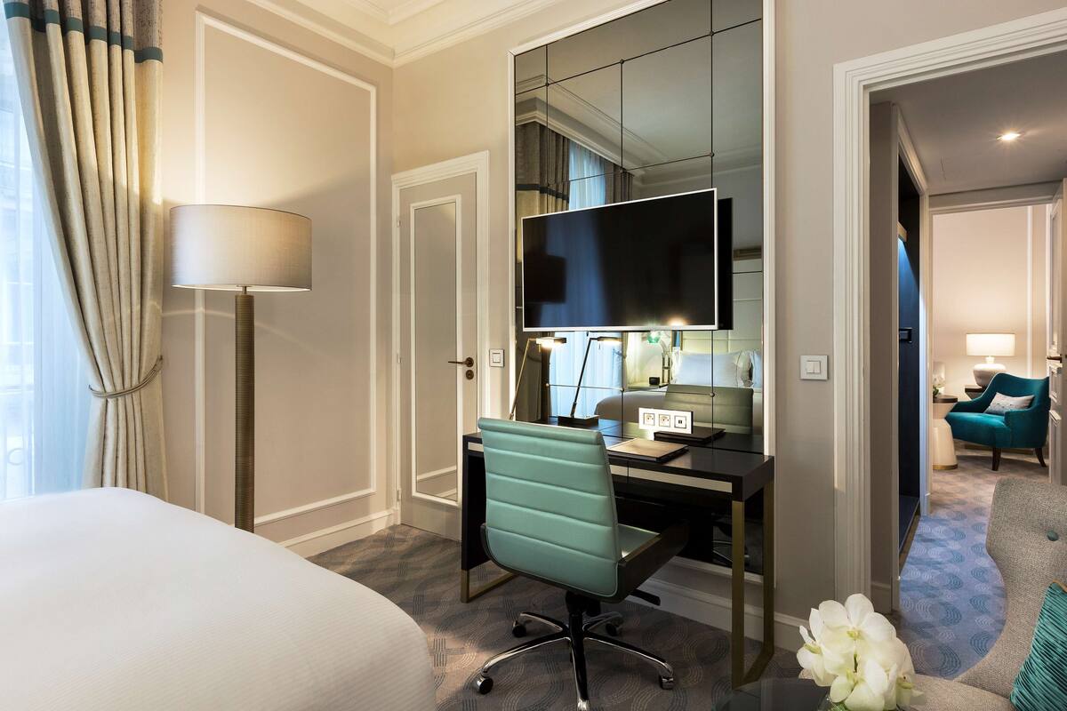 Habitación de hotel con cama, escritorio, silla, televisor y vista a otra habitación.