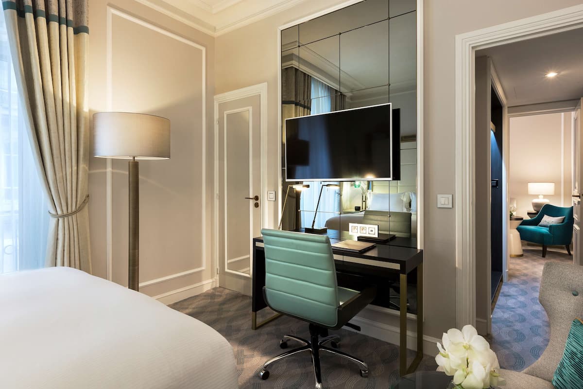 A hotel room with a bed, desk, chair, TV, and a view into another room.