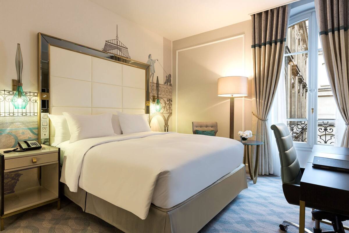 Habitación de hotel con una cama grande, un escritorio, una silla, una mesita de noche con teléfono, una lámpara y una ventana con cortinas.