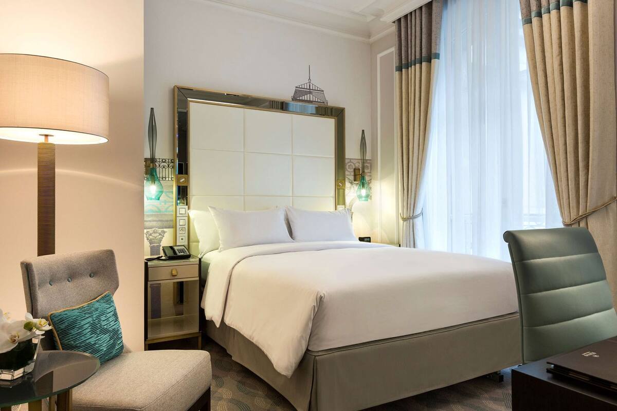 Habitación de hotel con cama, una silla, un escritorio, una lámpara y ventana con cortinas.
