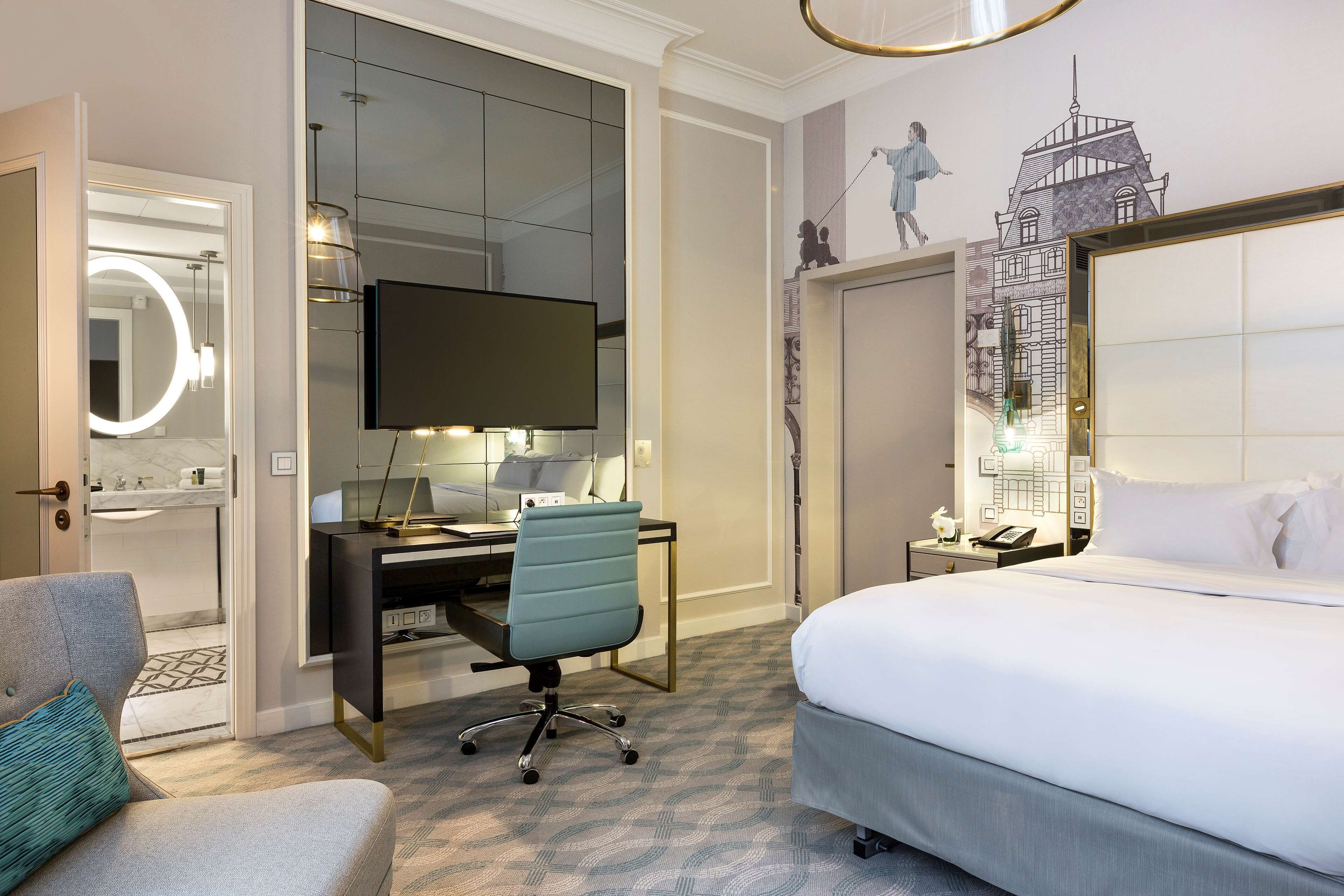 A hotel room with a bed, a desk with a computer, a chair, a TV, a mirror, and a wall with a cityscape mural.