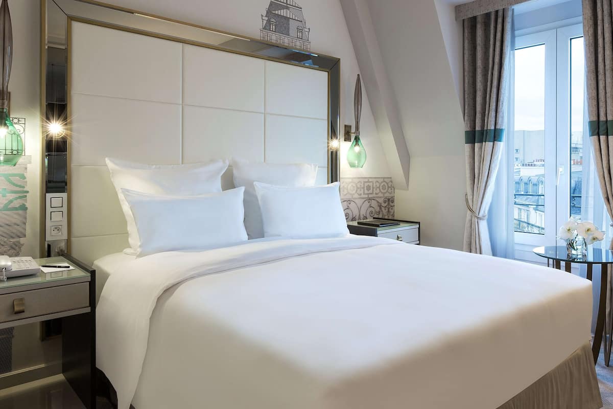Una cama bien hecha con sábanas blancas, una mesita de noche con lámpara y vistas de la ciudad a través de la ventana.