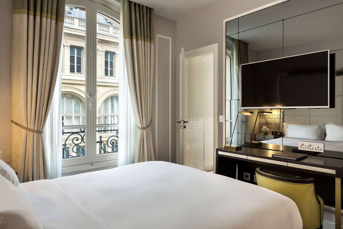 Habitación de hotel con cama, un escritorio con televisor de pantalla plana, un espejo y vista a un edificio a través de la ventana.