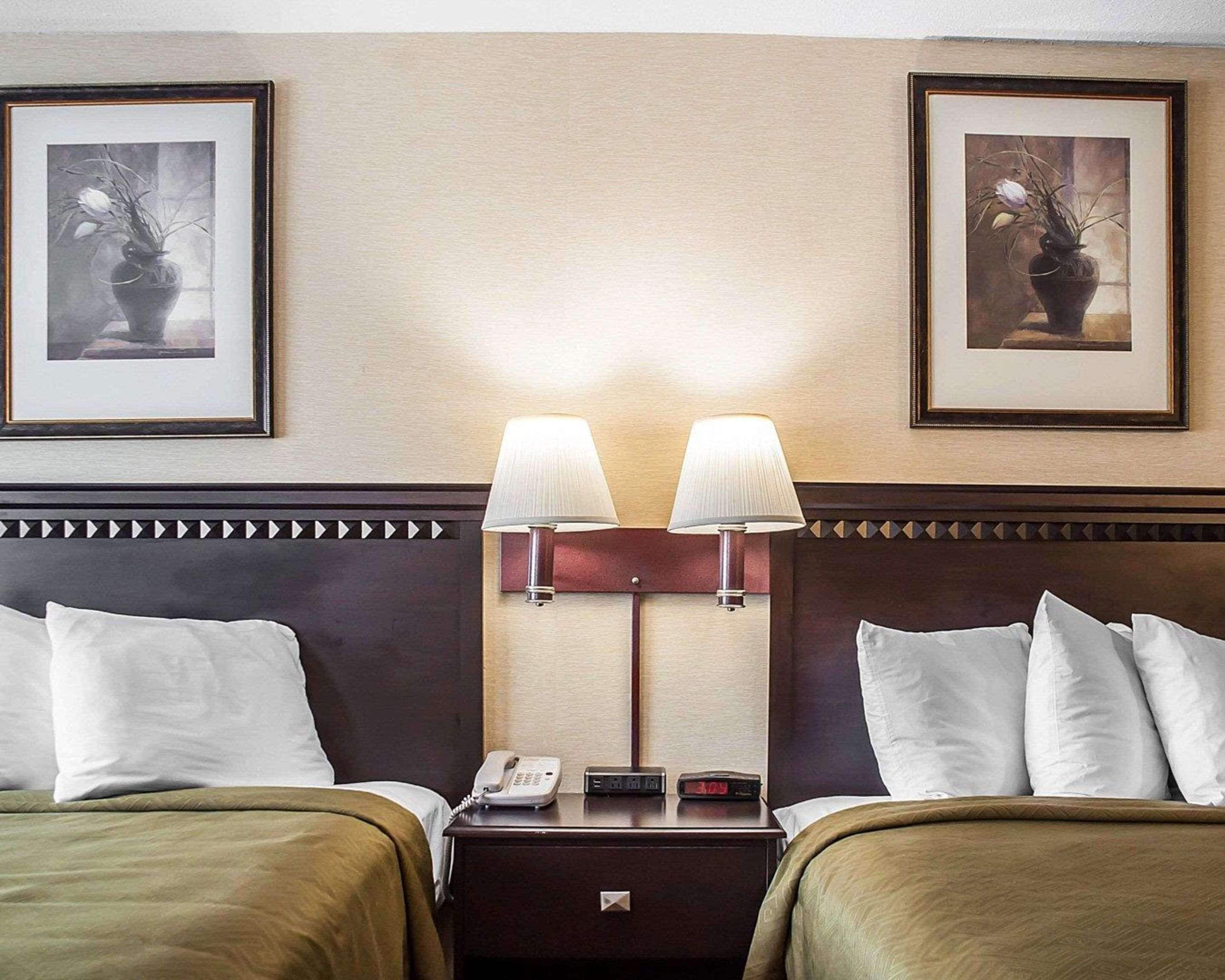 A hotel room with two beds, a nightstand with a phone and clock, and two wall-mounted lamps.