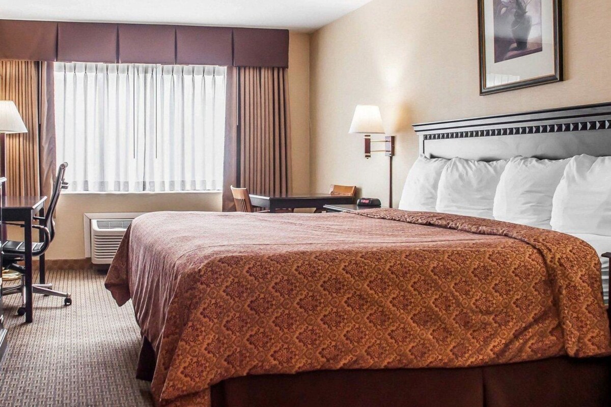 A hotel room with a large bed, a desk, a chair, a window with curtains, and a wall-mounted lamp.