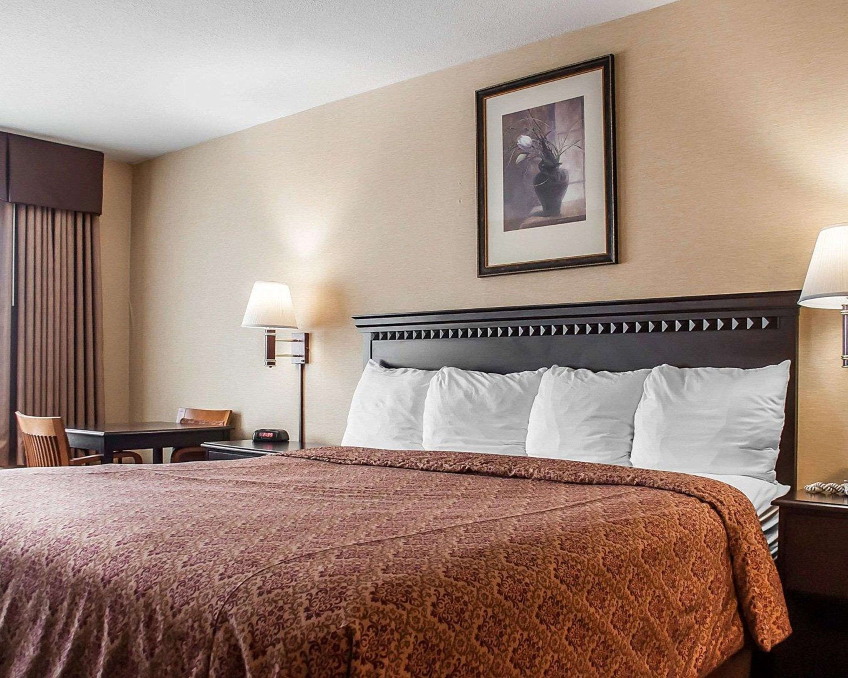 A hotel room with a large bed, two bedside lamps, a framed picture on the wall, and a small table with a chair.