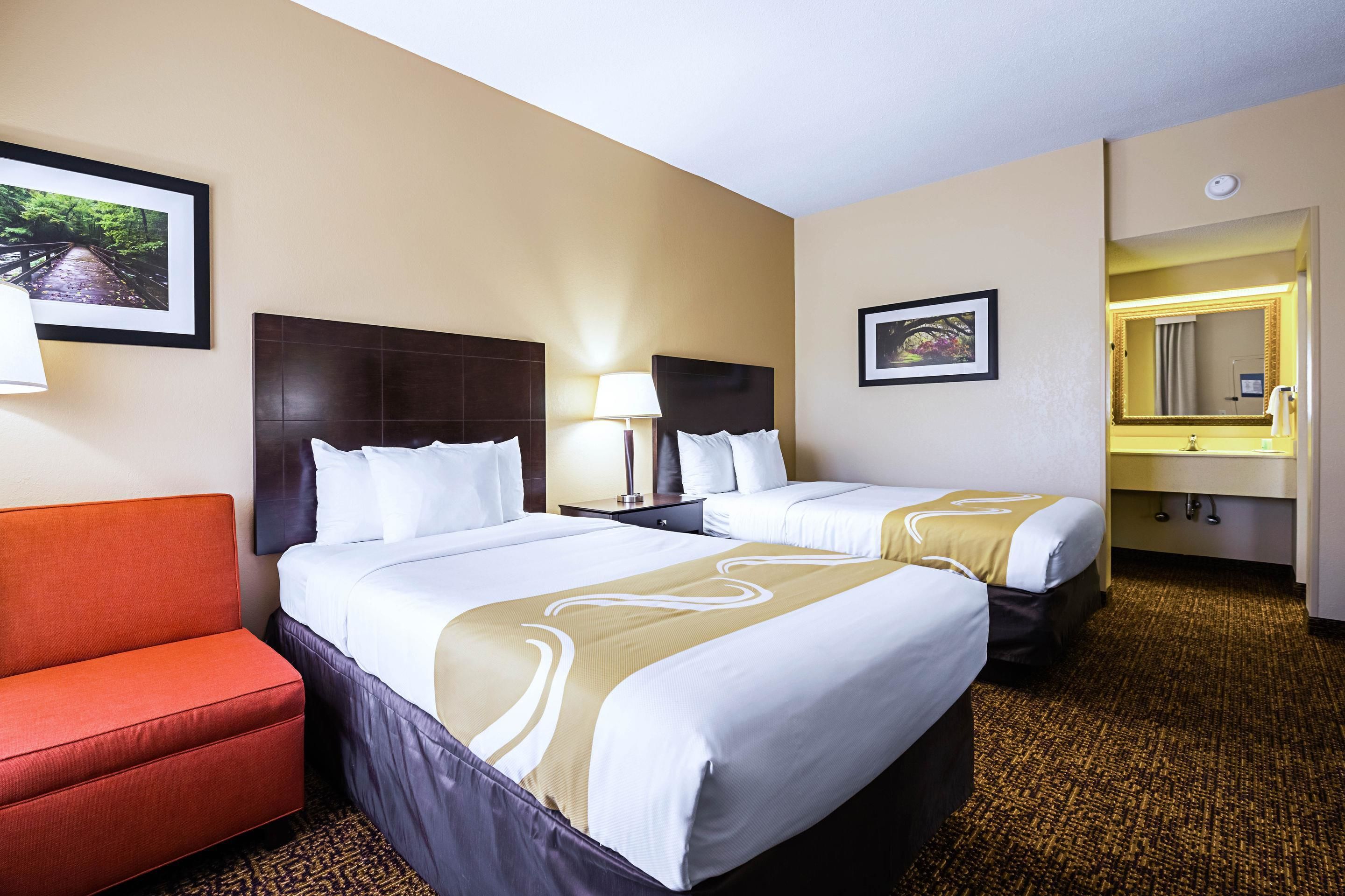 A hotel room with two beds, a red chair, a bathroom with a mirror, and framed pictures on the walls.
