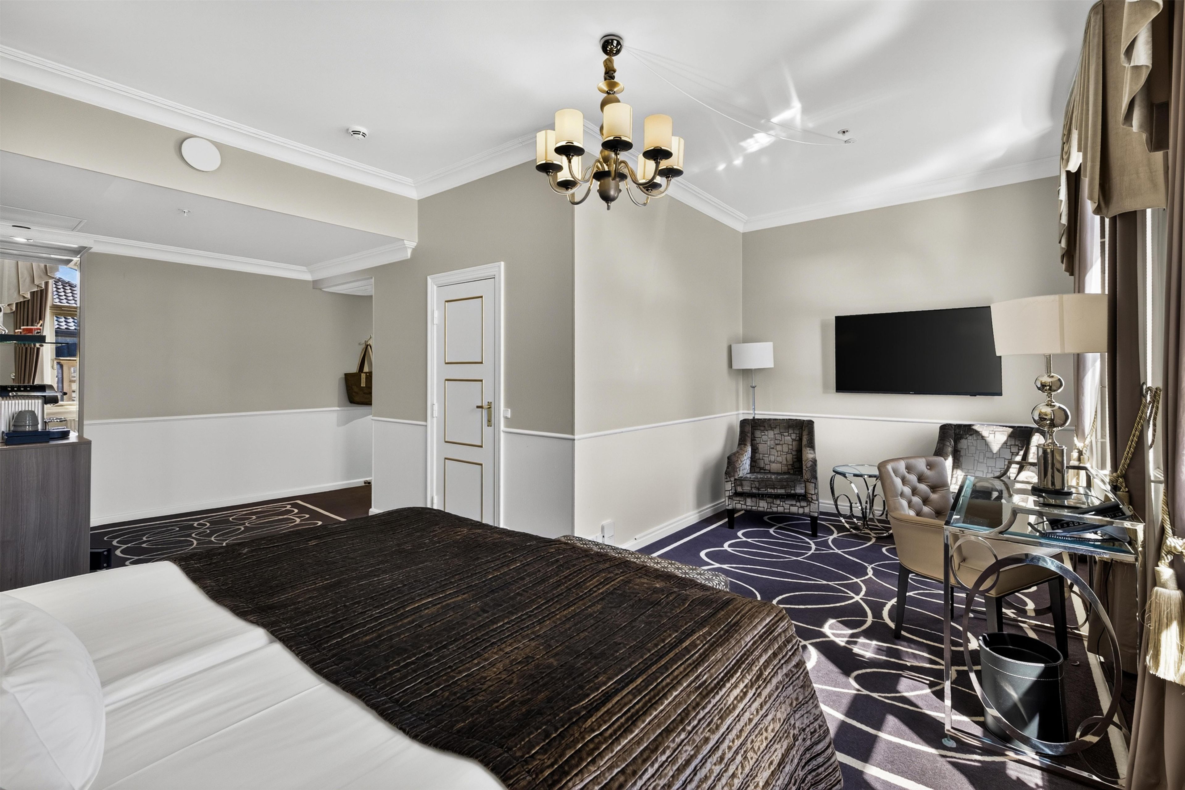 A hotel room with a large bed, a chandelier, a television, a seating area with a chair, and a desk with a lamp.